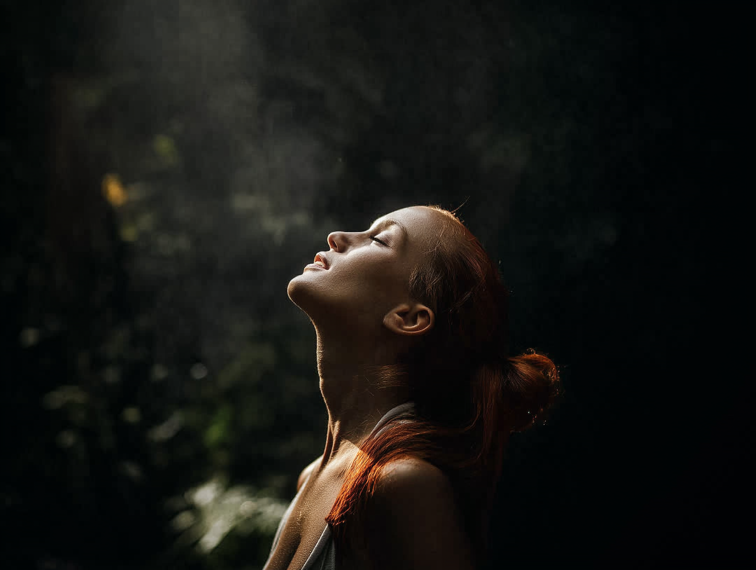 A woman with red hair and fair skin has her eyes closed and face tilted upward, basking in sunlight in a forest setting.