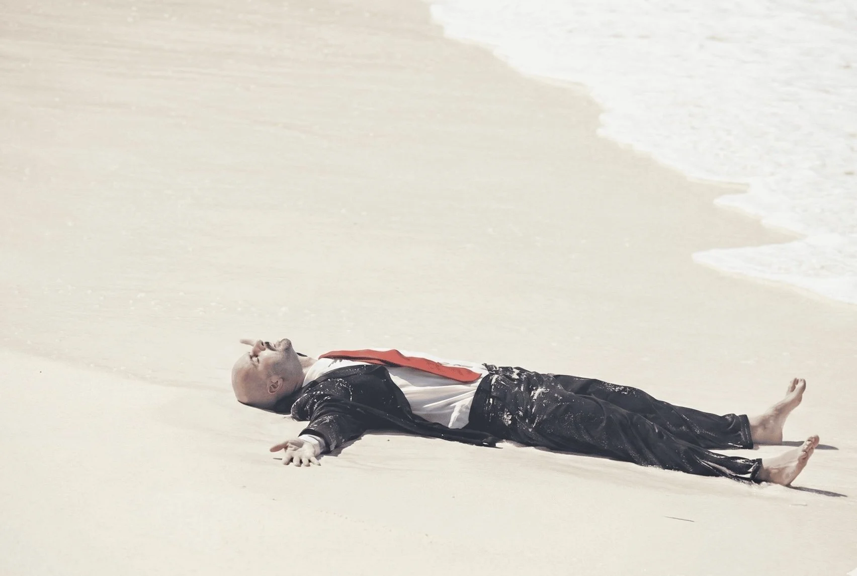 A man lying on a sandy beach near the water, dressed in a suit with a red tie, appearing to be unconscious or resting.