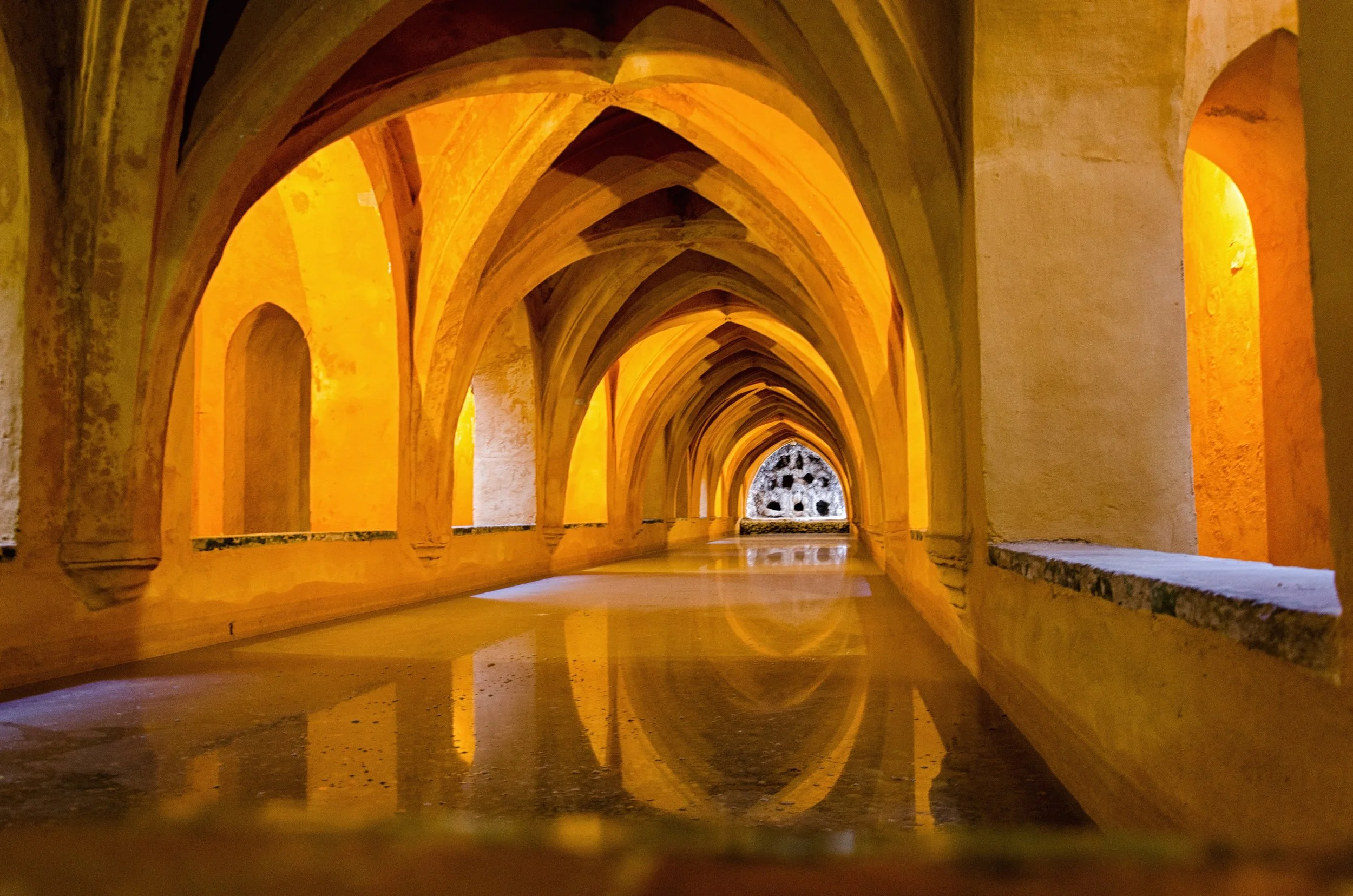 Interior view of a corridor with gothic arches illuminated by warm yellow lighting, with a reflective floor and a circular opening at the end.