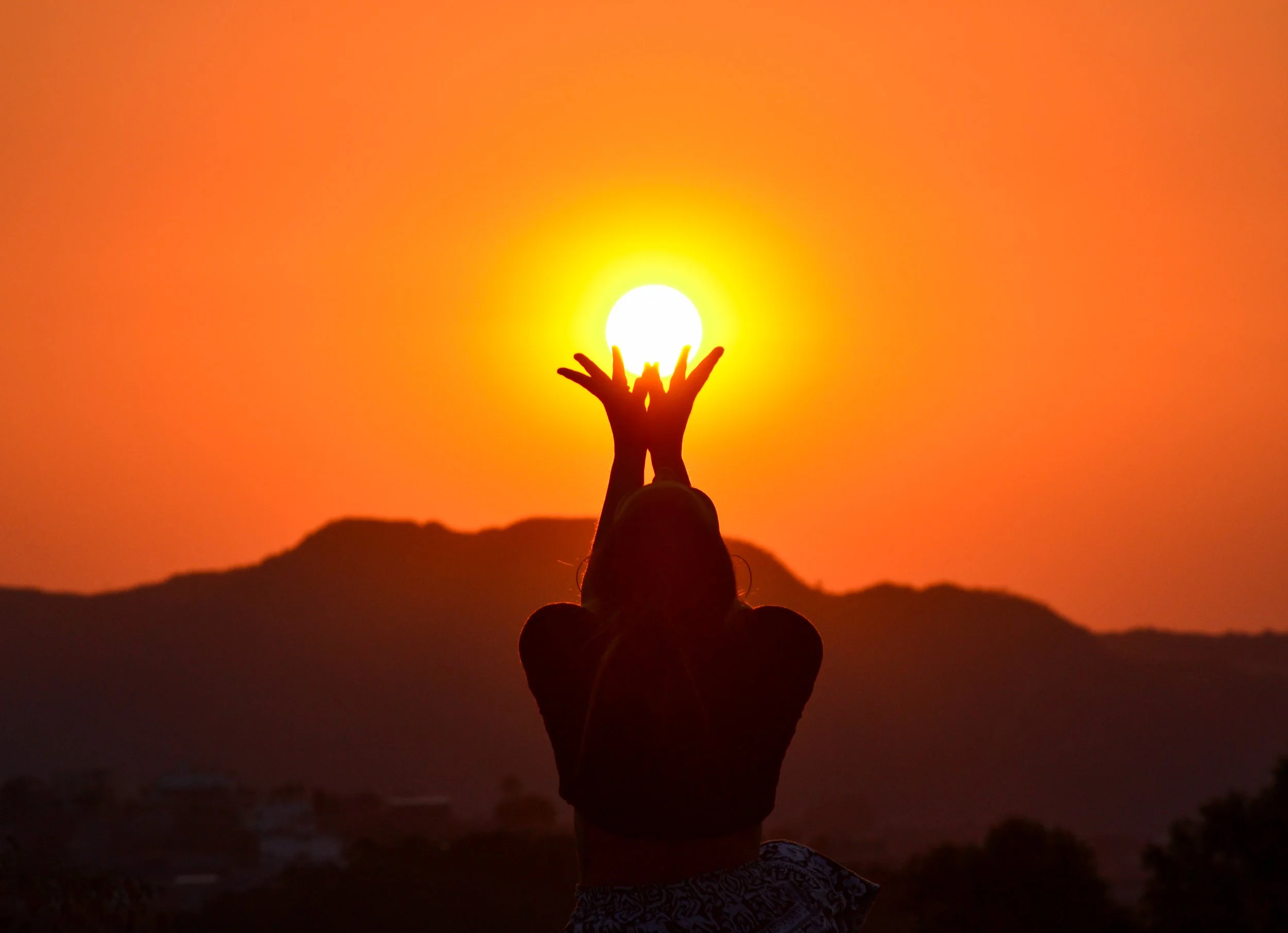 Silhouette of a person holding their hands up towards the setting sun during sunset with mountains in the background.