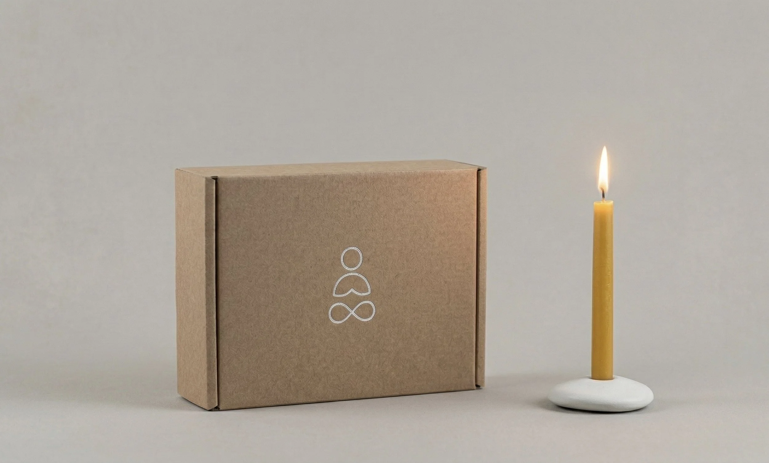 The packaging of The Loop meditation candle with meditationawhite logo on the front, beside a yellow candle burning in a ceramic holder, on a plain neutral background.