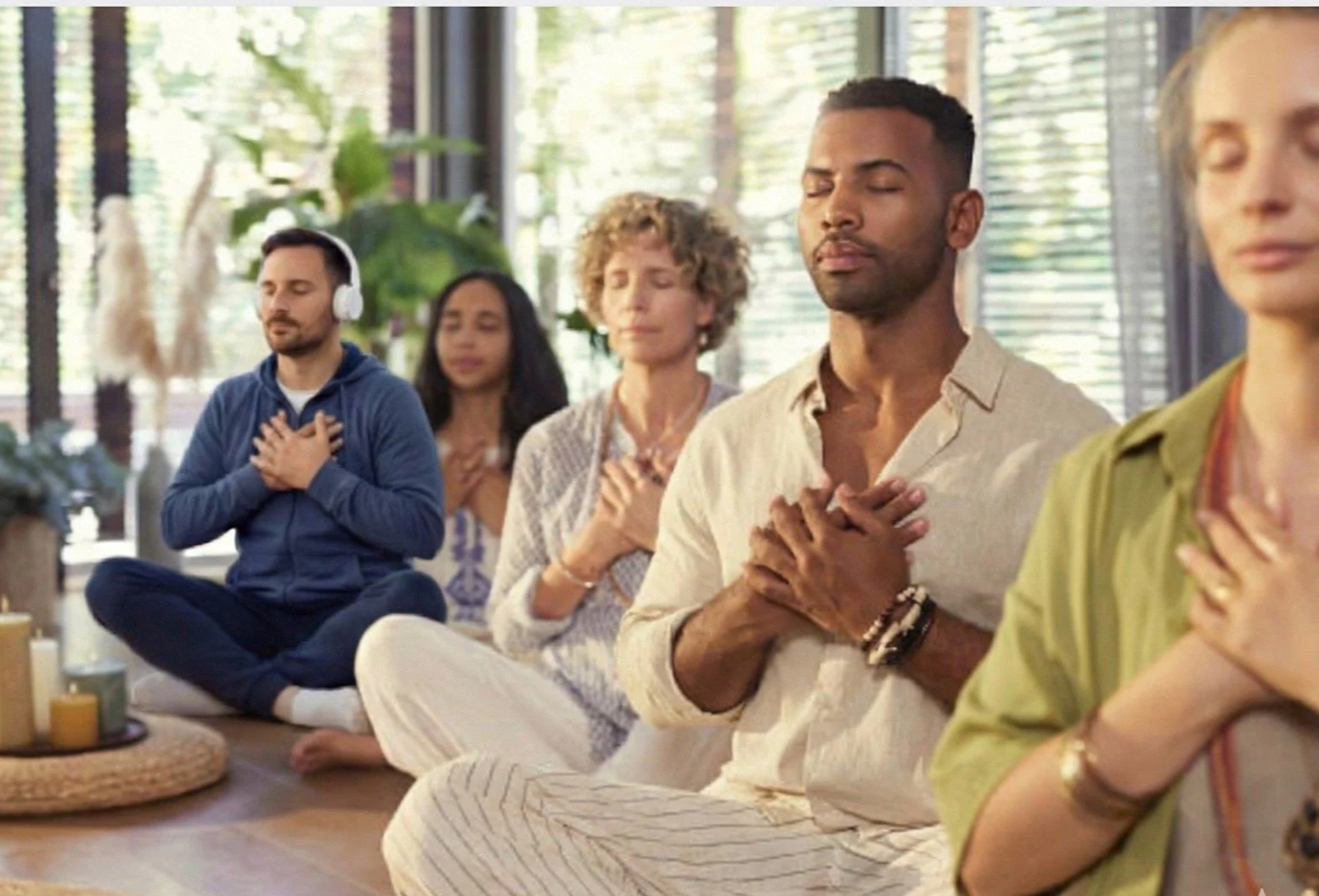 Group of individuals practicing meditation and mindfulness in a serene indoor space, sitting cross-legged with hands over their hearts, eyes closed.