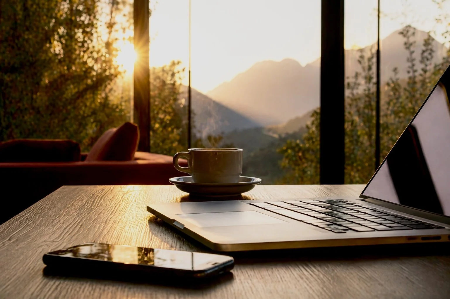 A laptop, a smartphone, and a cup on a wooden table with a view of mountains and trees through large windows at sunset.