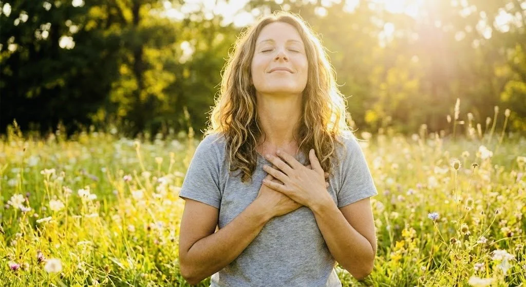 Woman with closed eyes and hand on chest standing in a flower field during sunlight.