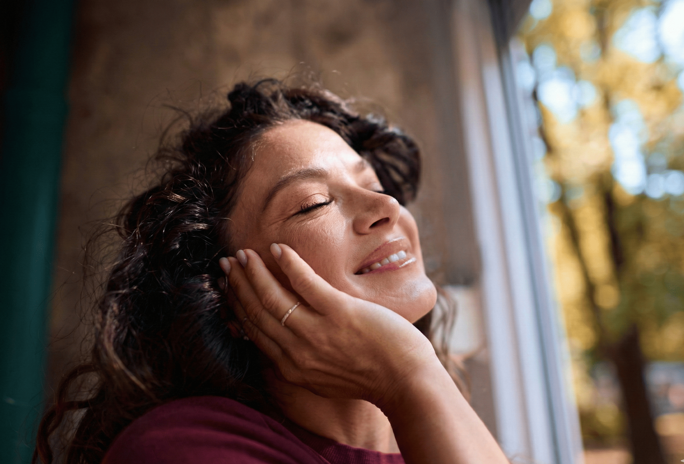 A woman with curly dark hair smiling and relaxing with her eyes closed, resting her face on her hand near a window with trees outside. She represents  quiet embodied  joy.