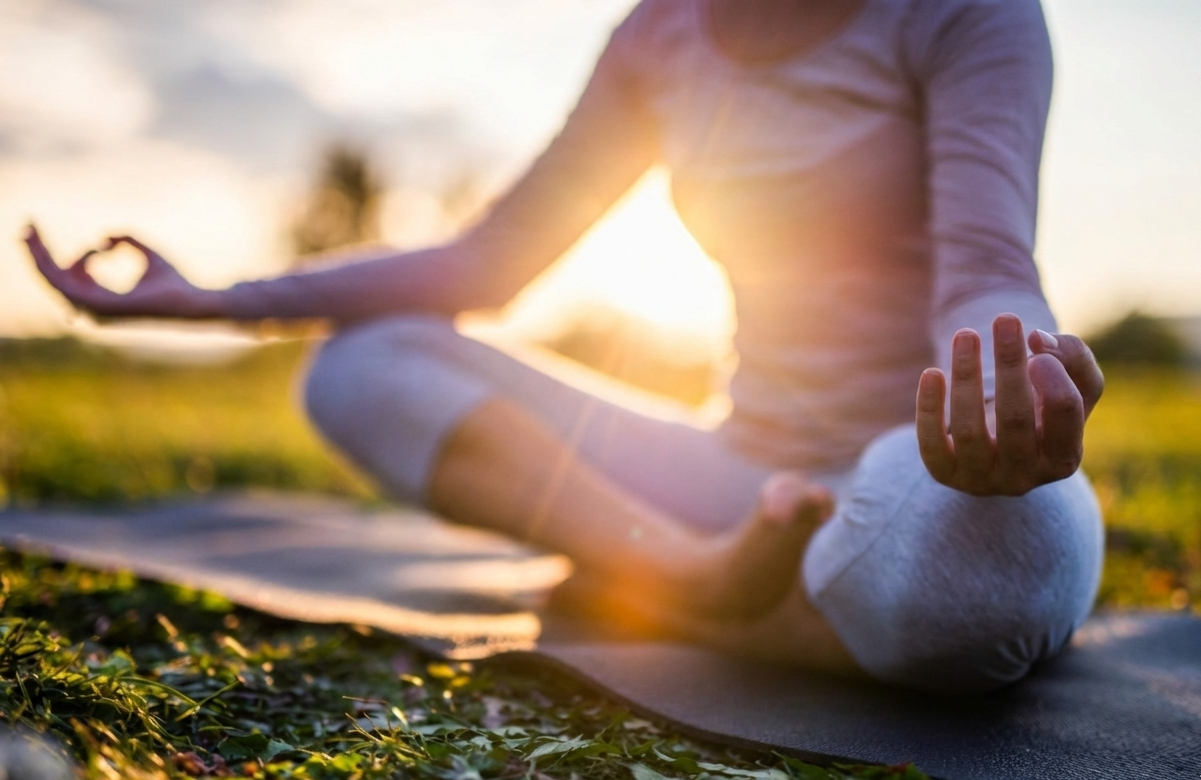 Person practicing yoga outdoors during sunset, sitting cross-legged on a yoga mat with hands resting on knees and fingers forming mudra. Connecting to the peace energy.