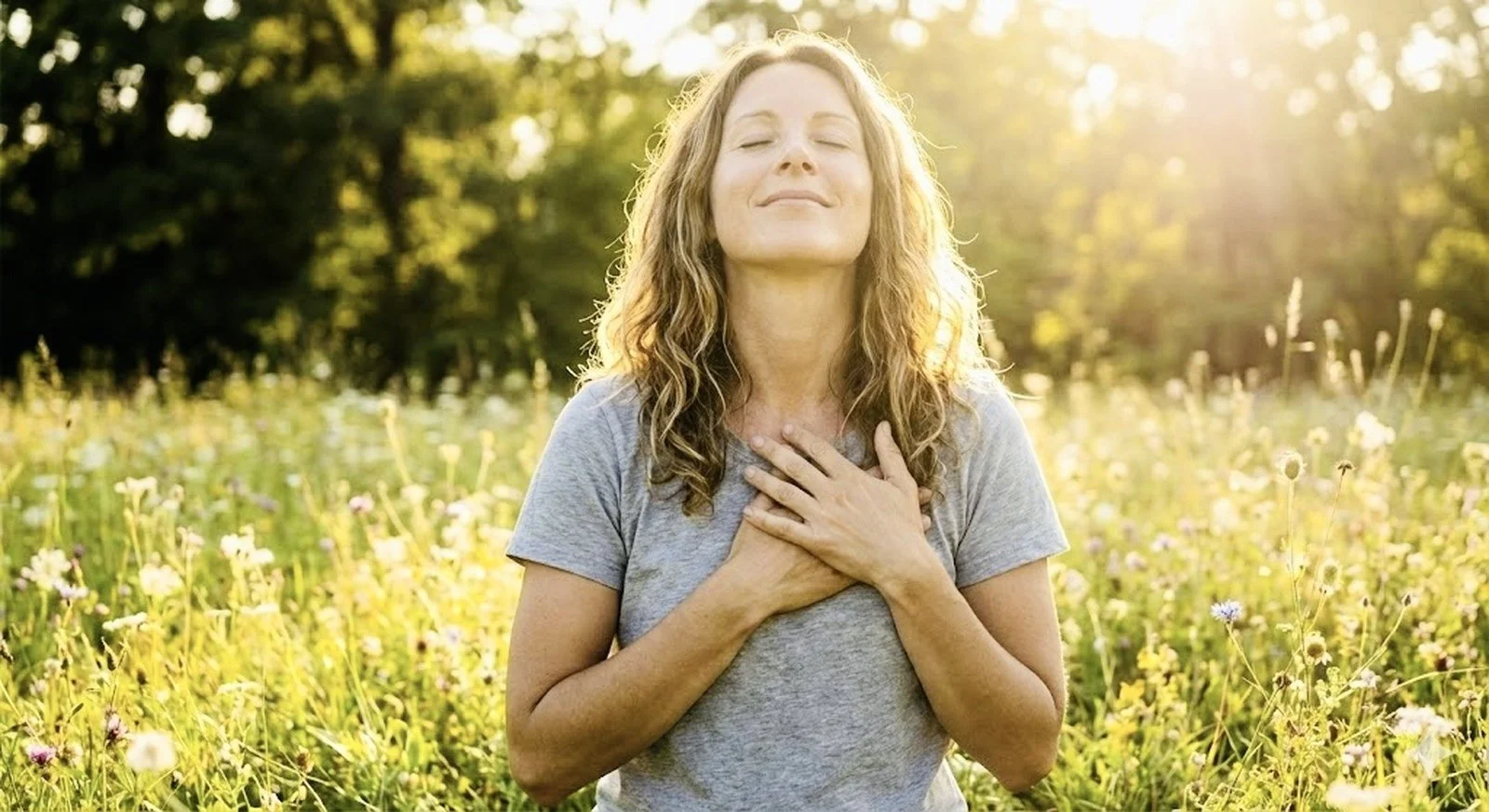 Woman with closed eyes and hand on heart chakra standing in a flower field during sunlight.
