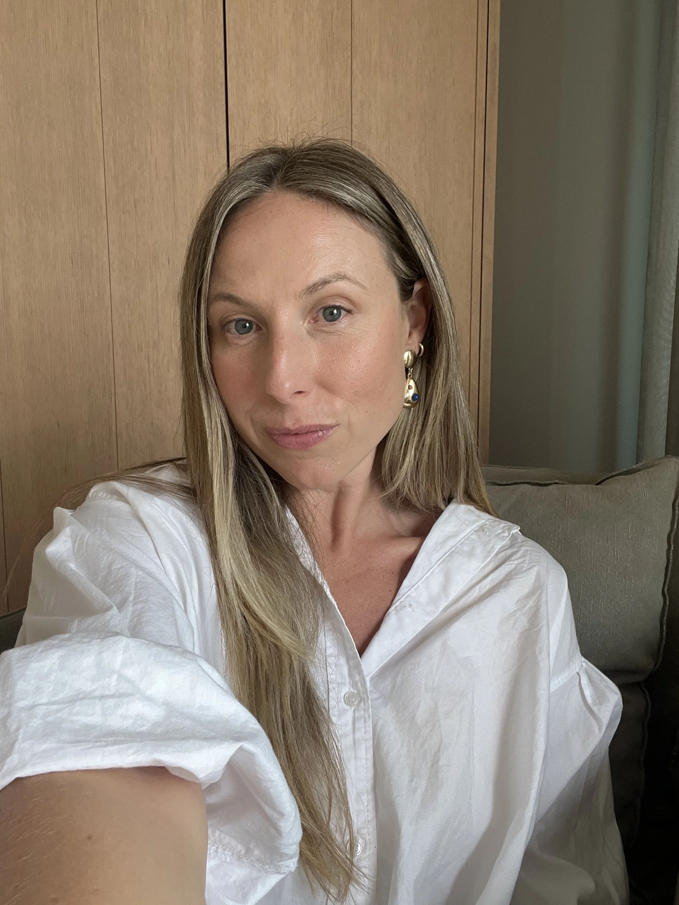 Close-up of a woman with long blonde hair wearing a white button-down shirt and gold earrings, sitting on a beige couch with a wooden wall in the background.