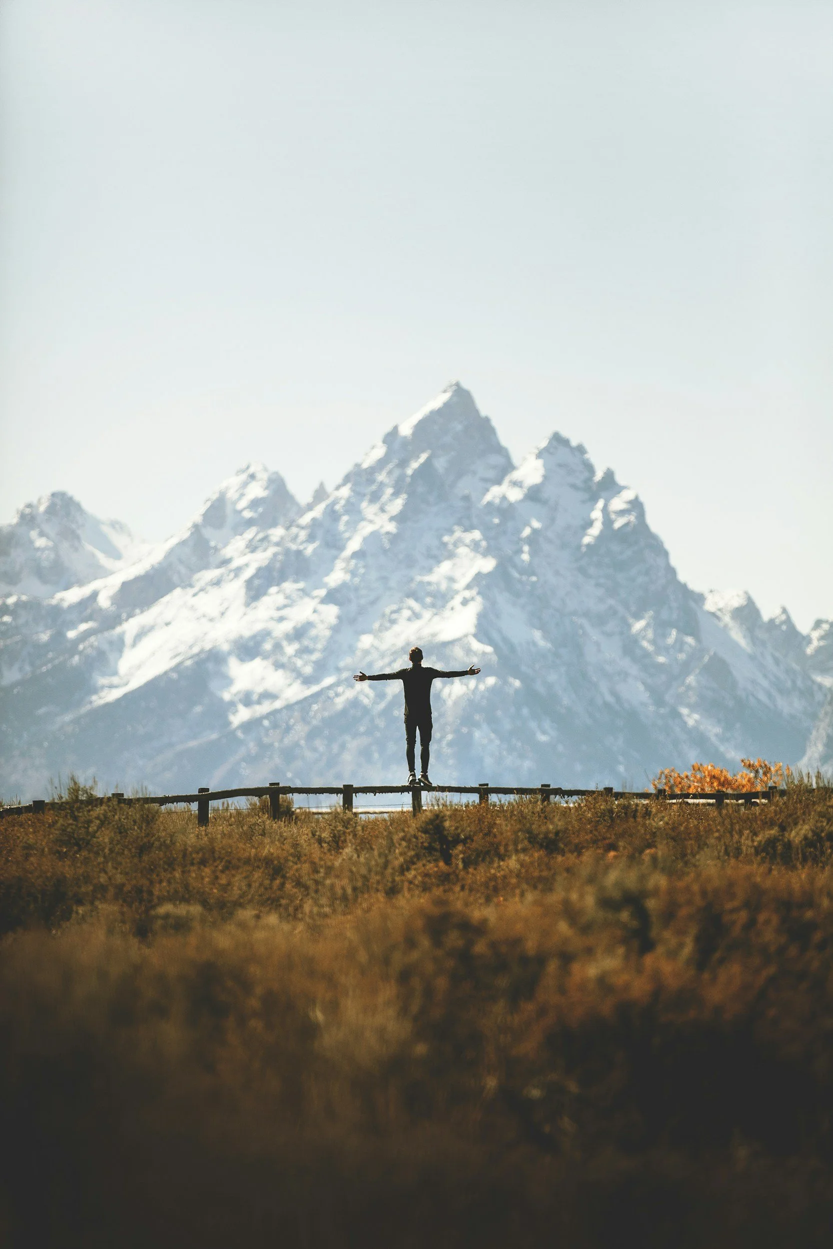 Person standing on a wooden fence with arms outstretched, overlooking a mountain range with snow-capped peaks.