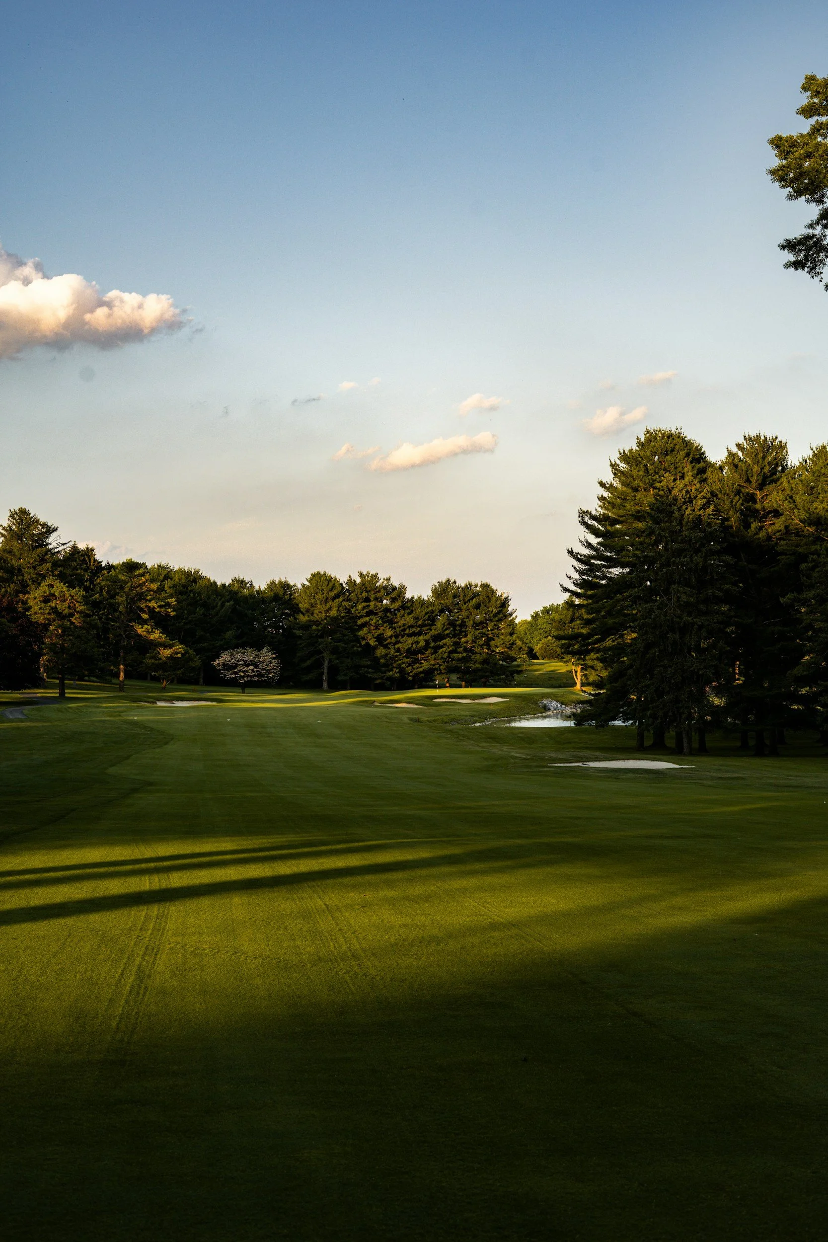 A golf course with green grass, trees, a water hazard, and a partly cloudy sky in the background during daytime.
