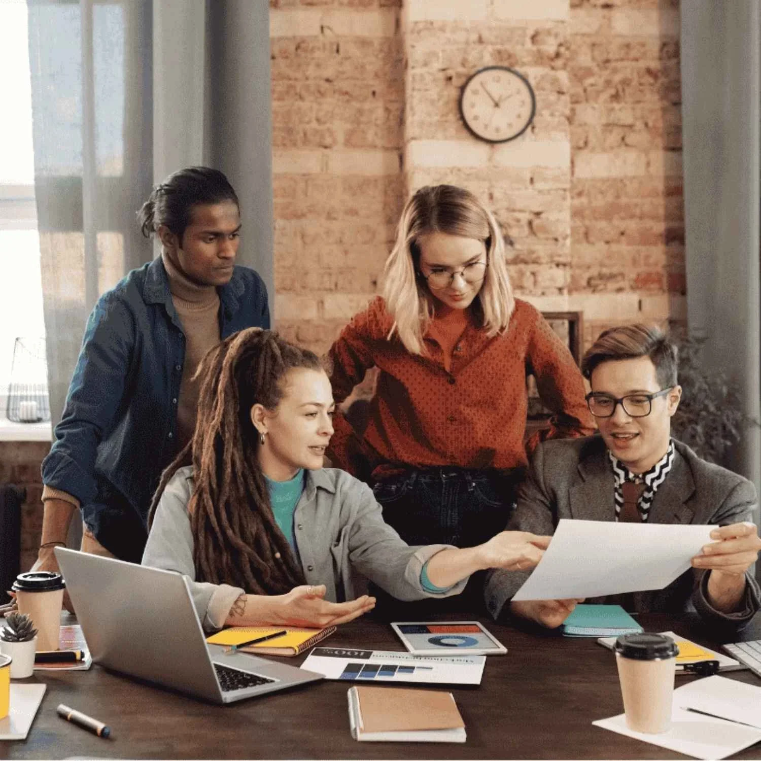 Five people working together around a table in a modern office with exposed brick walls. One woman with dreadlocks is showing a document to a man in glasses. Two other women, one with glasses and one with blonde hair, are observing. The table has laptops, notebooks, coffee cups, and various papers.