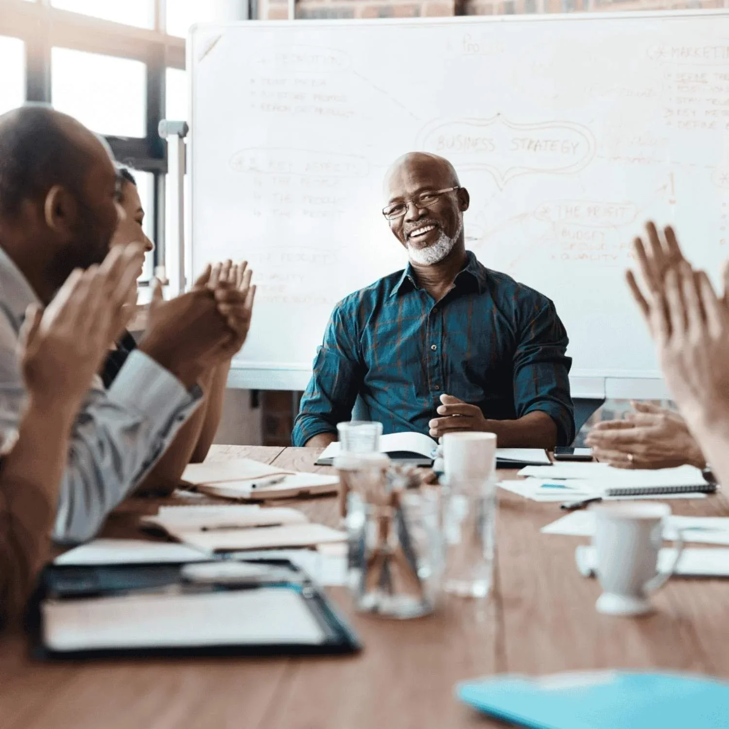 A group of diverse people in a business meeting, clapping and smiling, with a man in a blue shirt leading the discussion in front of a whiteboard.