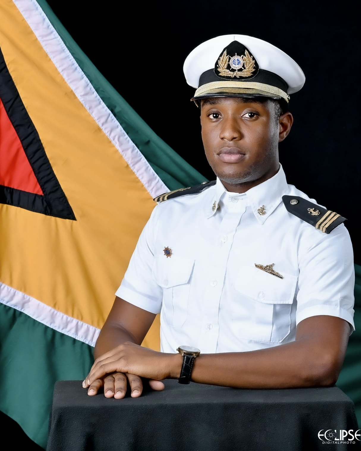 Young man in a white military uniform with a captain's insignia, standing in front of a flag with green, yellow, black, white, red, and black colors, against a black background.
