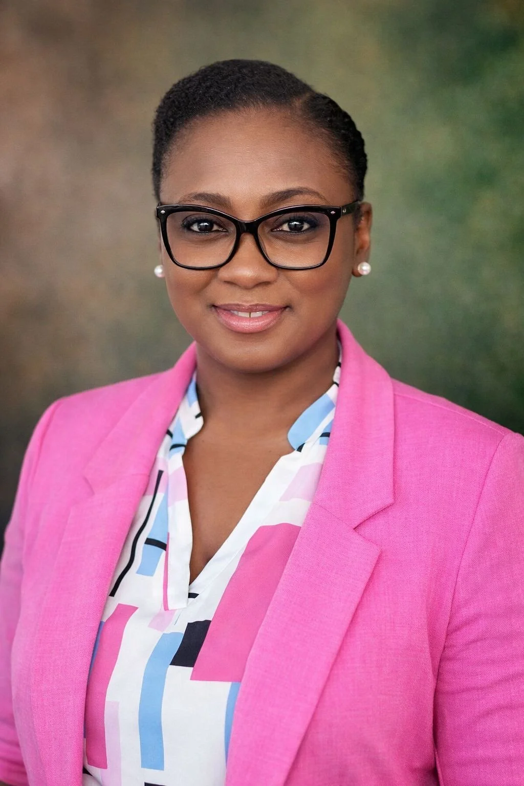 Dr. Kamecia Blake, glasses, a pink blazer, and a patterned blouse, smiling, with a blurred green background.