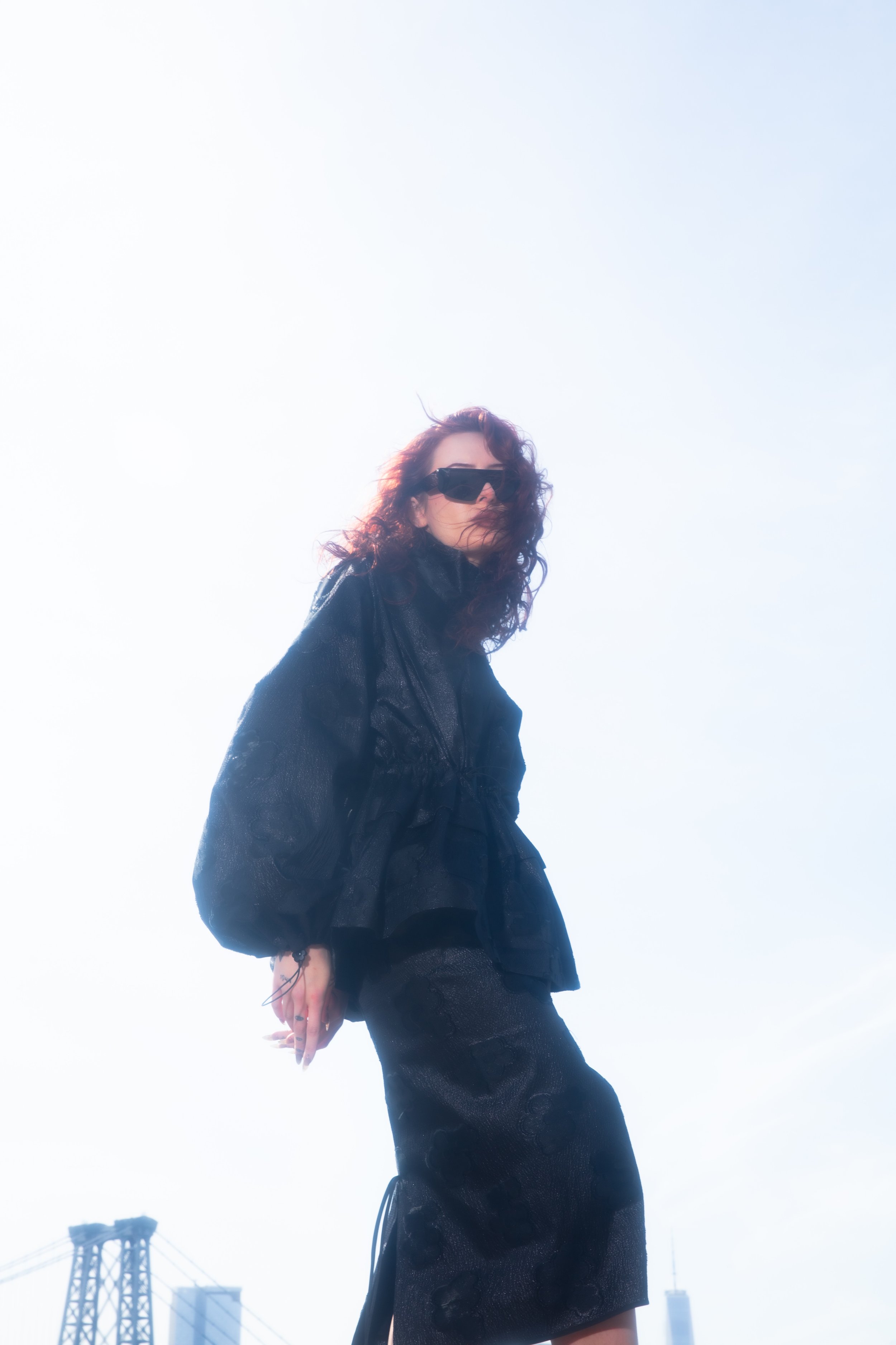 A woman with curly dark hair wearing black sunglasses, a black jacket, and a black skirt standing outdoors with the city skyline and a bridge in the background.