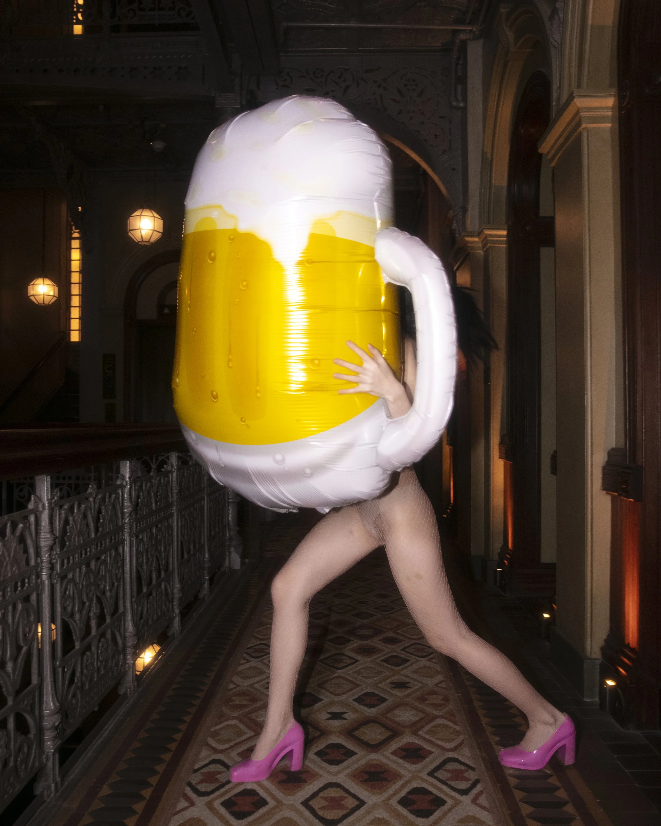 Person dressed as a large beer mug with foam on top, wearing pink high heels, standing in a historic building corridor.