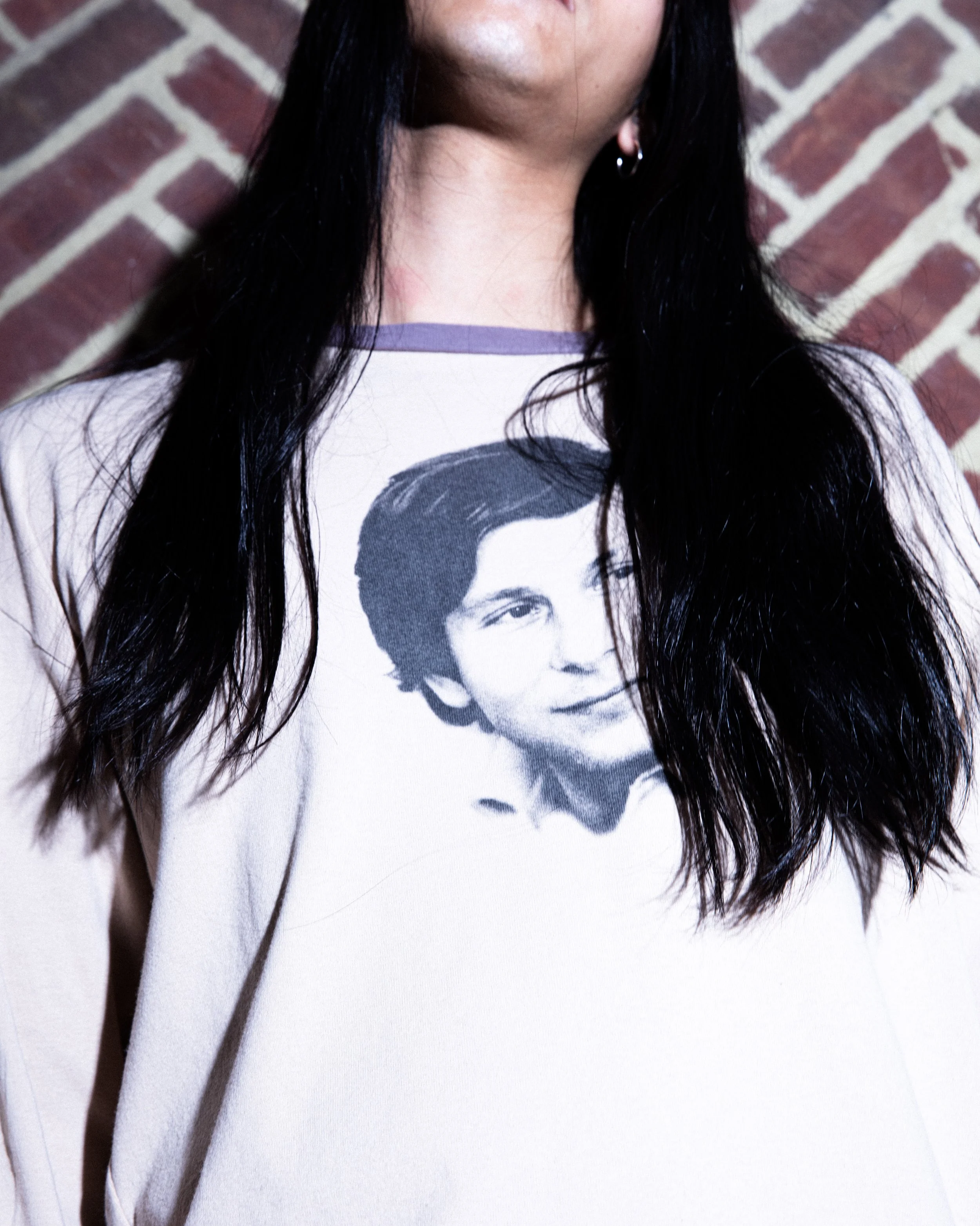 Person with long dark hair wearing a white shirt with a vintage photo of a woman printed on it, standing in front of a brick wall.