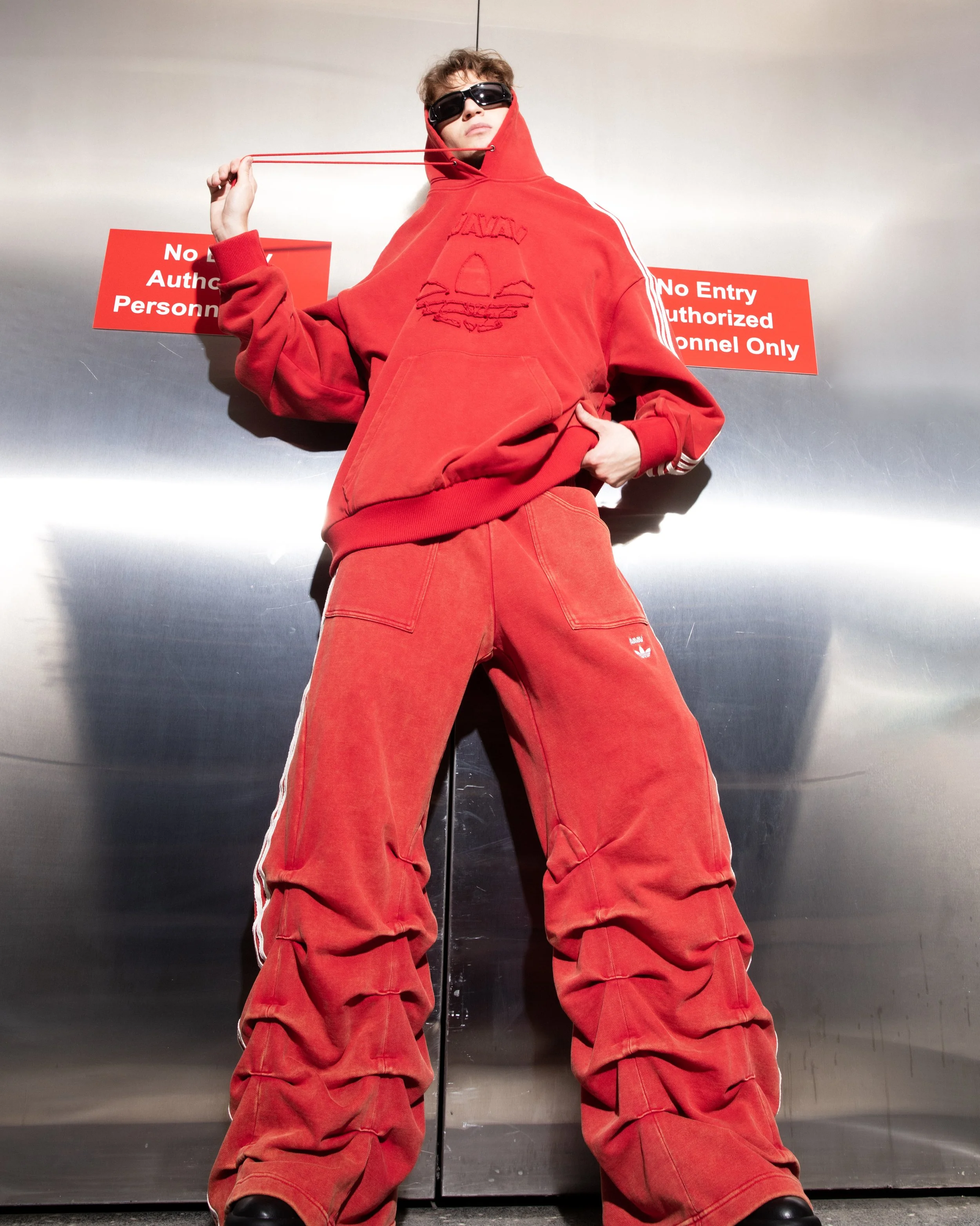 Person wearing a red hoodie, red pants, and sunglasses, standing in front of signs that read 'No Entry Authorized Personnel Only' in an industrial setting.
