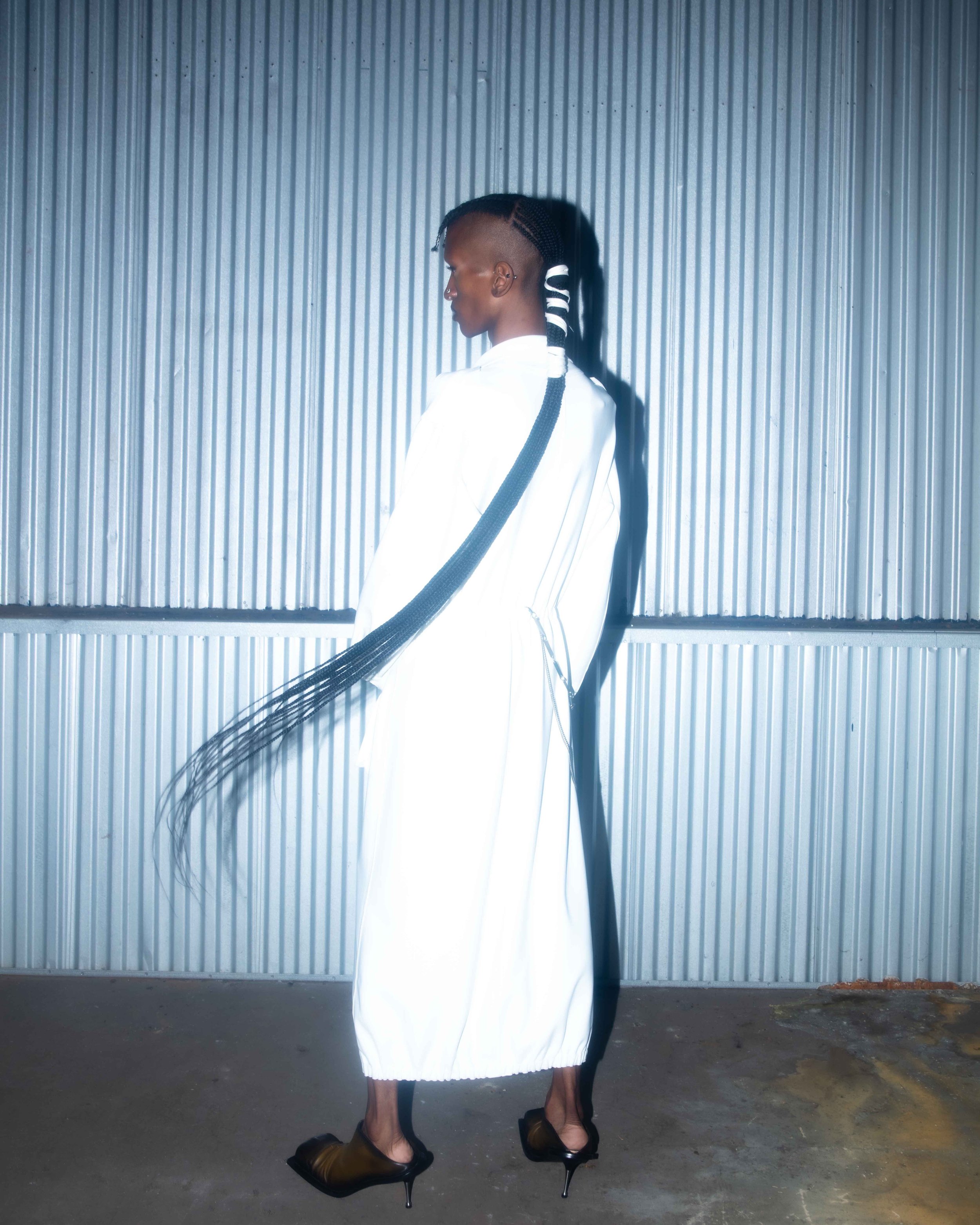 A woman in a white outfit with long braided hair standing in front of a corrugated metal wall.