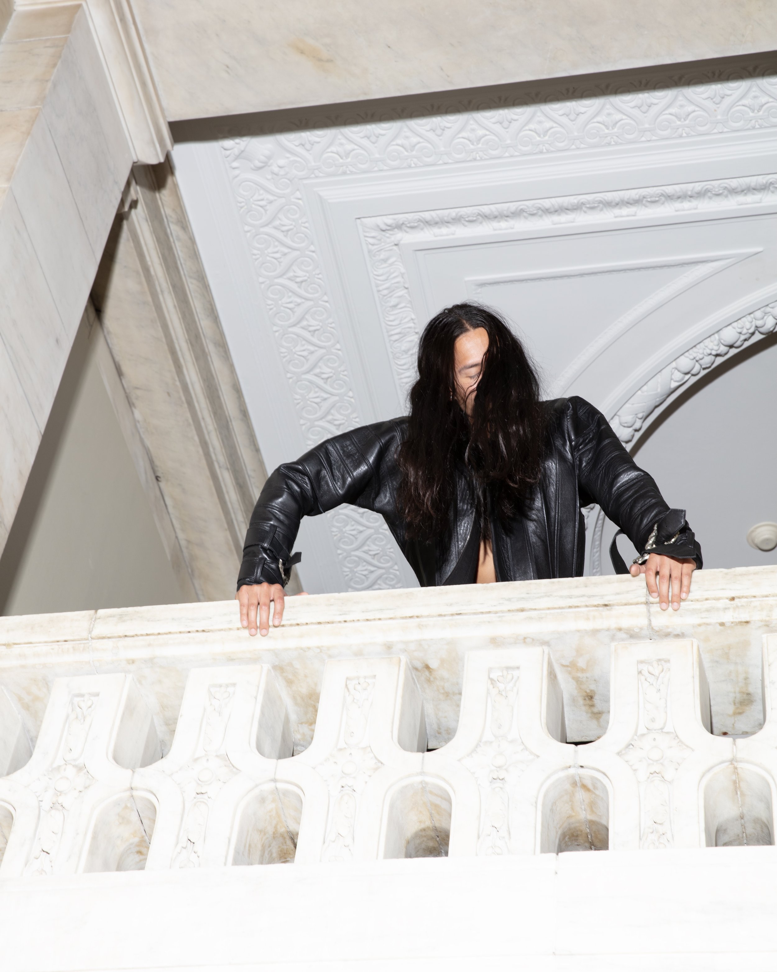 Person with long dark hair wearing a black leather jacket leaning over a decorative white marble balcony railing inside a building with ornate white ceiling