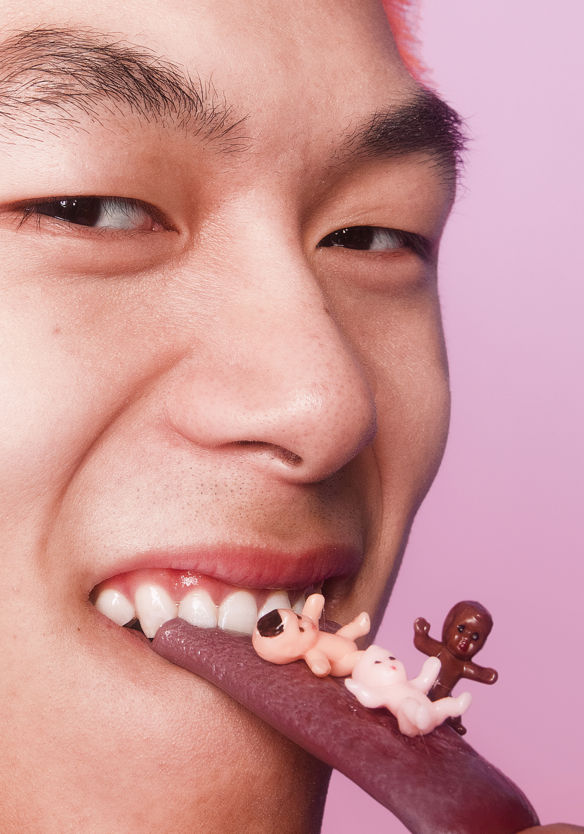 A person with glasses sticking out their tongue, which has three miniature gummy baby figures on it, including a pink baby on its back, a white baby, and a brown baby.