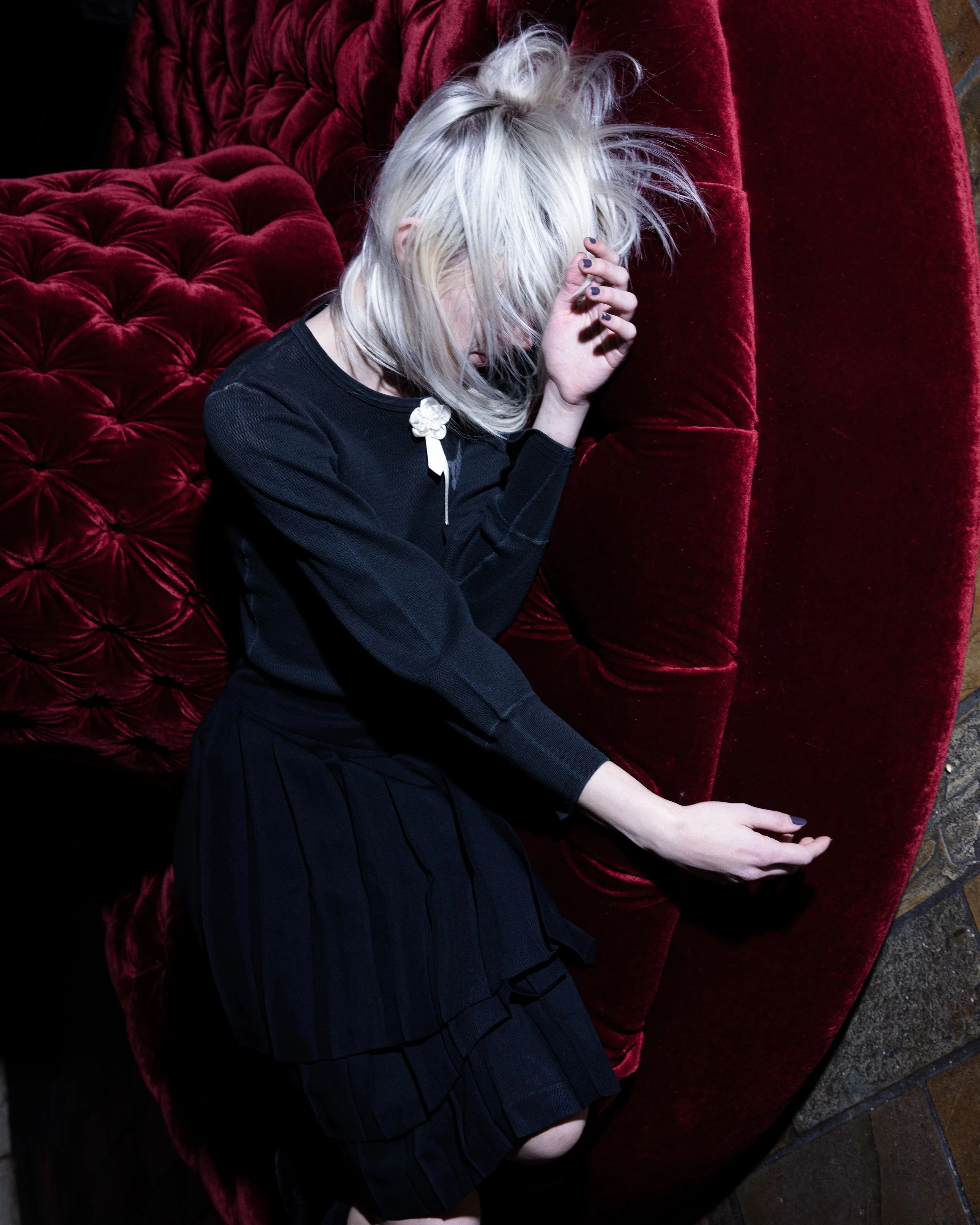 A woman with platinum blonde hair wearing a black dress, sitting on a red velvet sofa, holding her forehead with her left hand and extending her right arm.