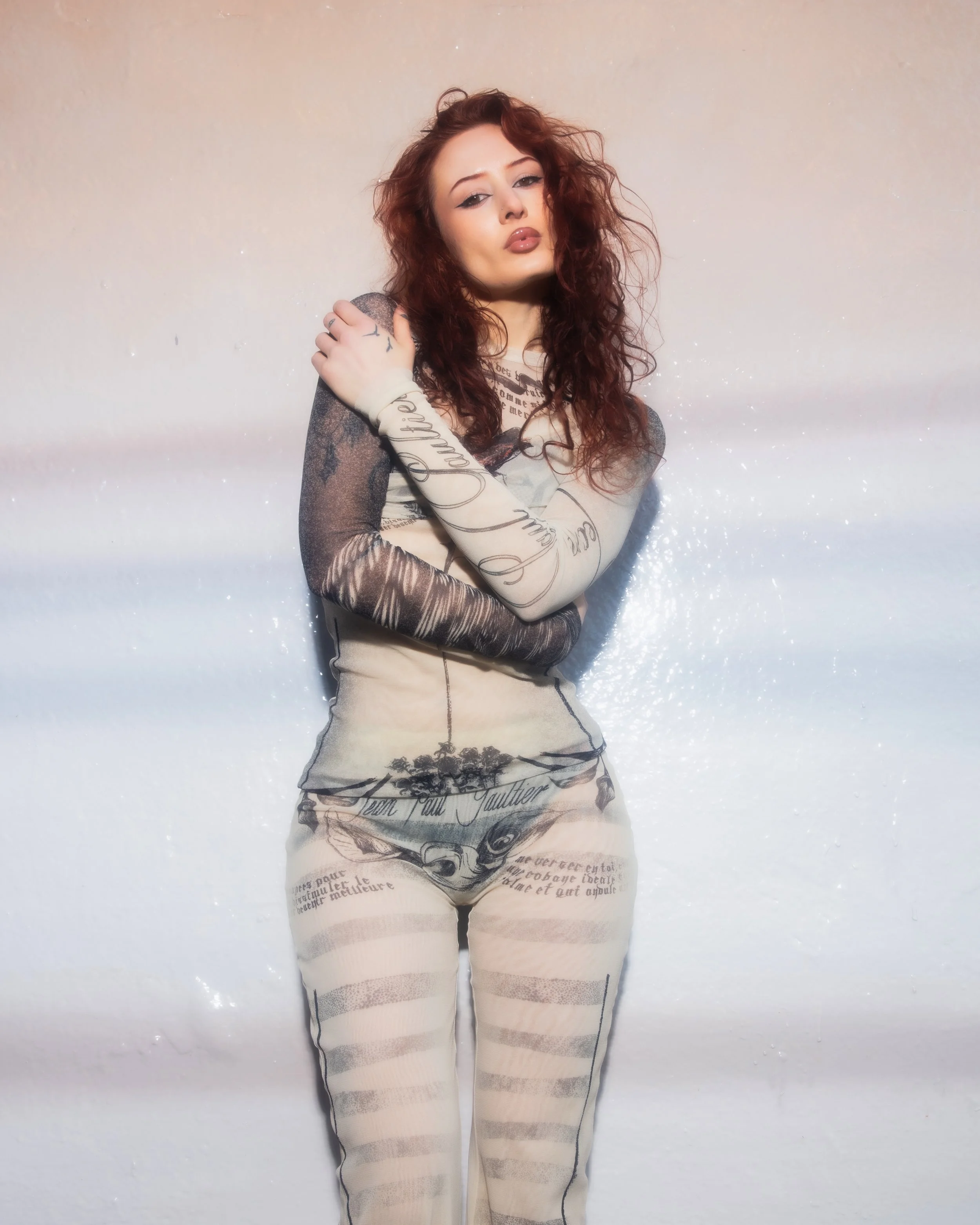 A woman with long curly red hair poses against a gradient background, wearing sheer clothing with printed text and images, and looking at the camera.
