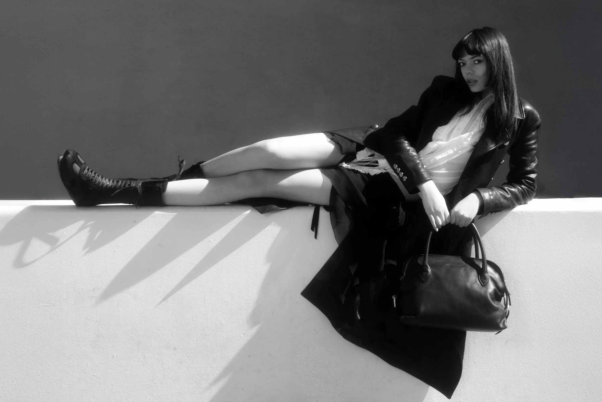 A black-and-white photo of a young woman lying on a wall, wearing a leather jacket, a skirt, and combat boots, holding a large handbag, with her eyes looking at the camera.