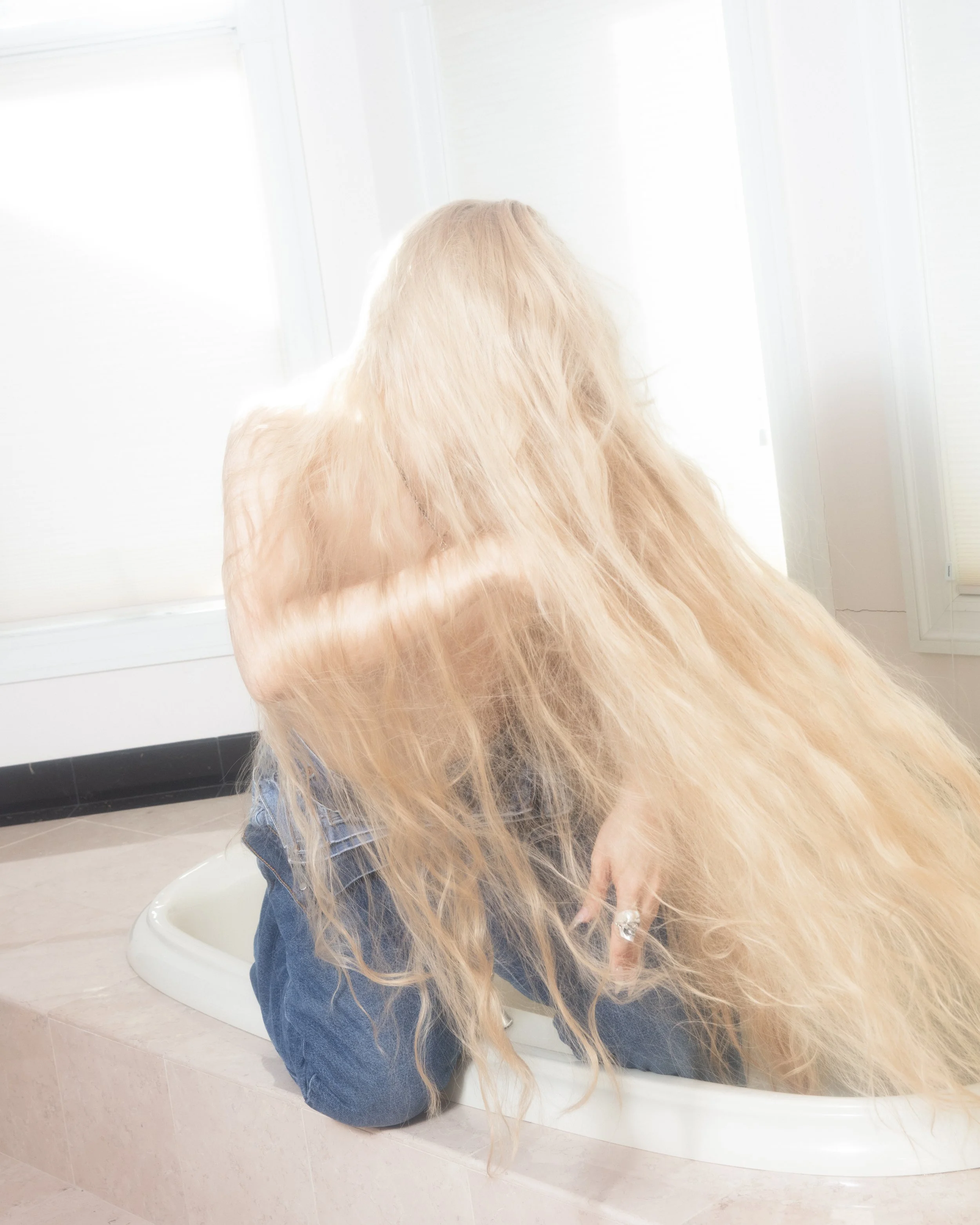 Person with long, curly, blonde hair sitting in a bathtub, facing away from the camera near a bright window.