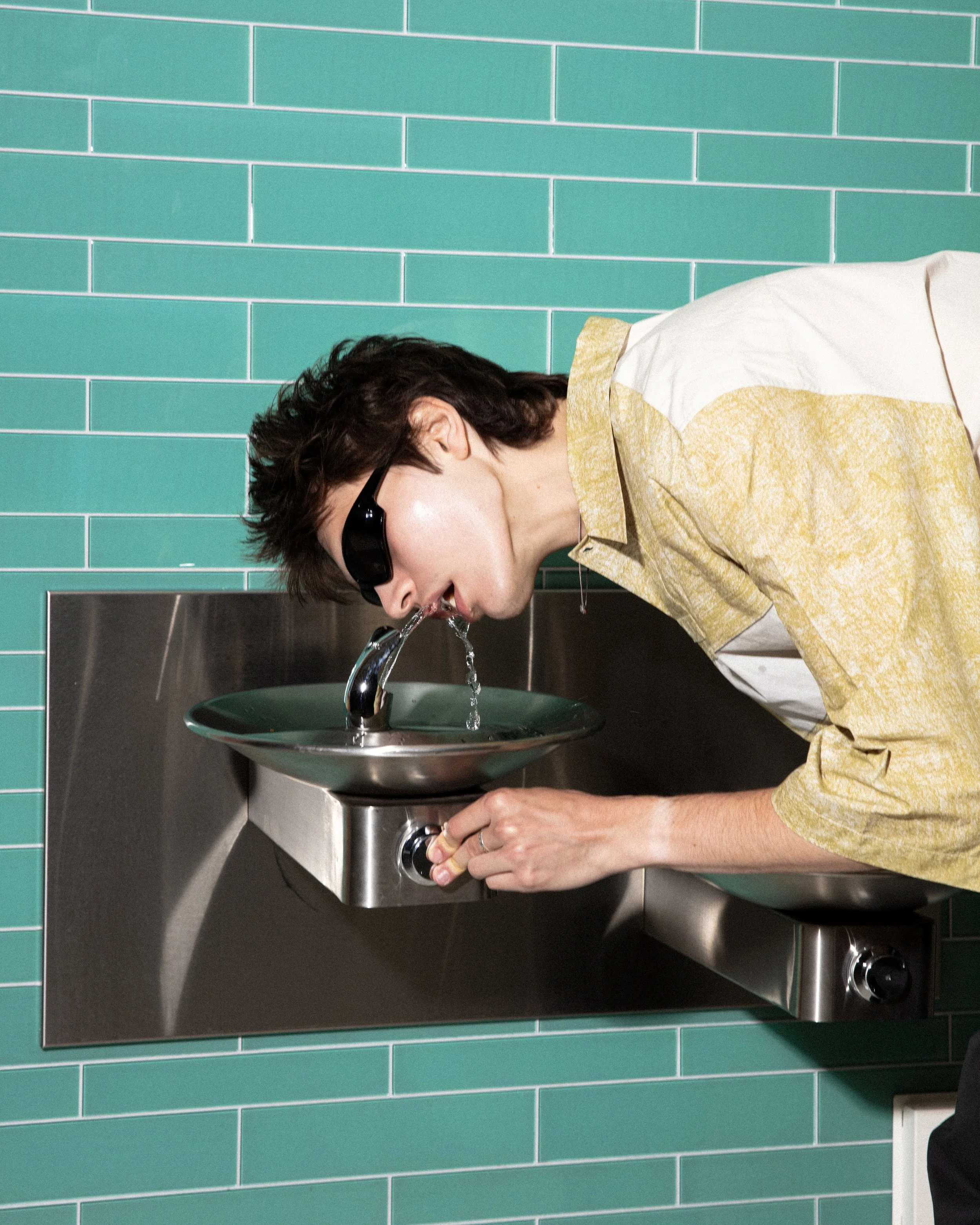 Person wearing sunglasses leaning over a drinking fountain, drinking water.