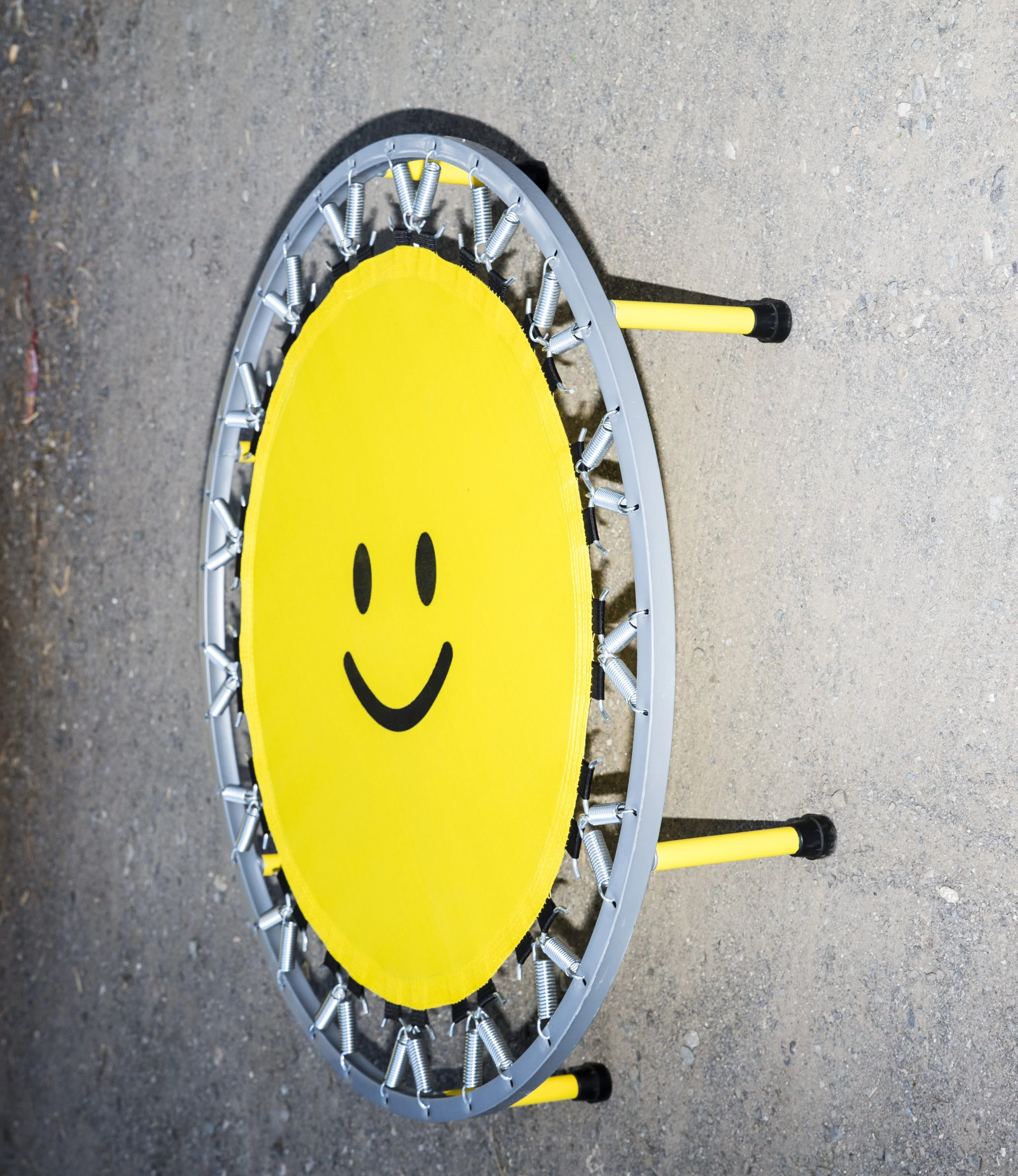Round mini trampoline with yellow surface featuring a black smiley face. The trampoline has metal springs and a gray frame, and it stands on a gravel surface.
