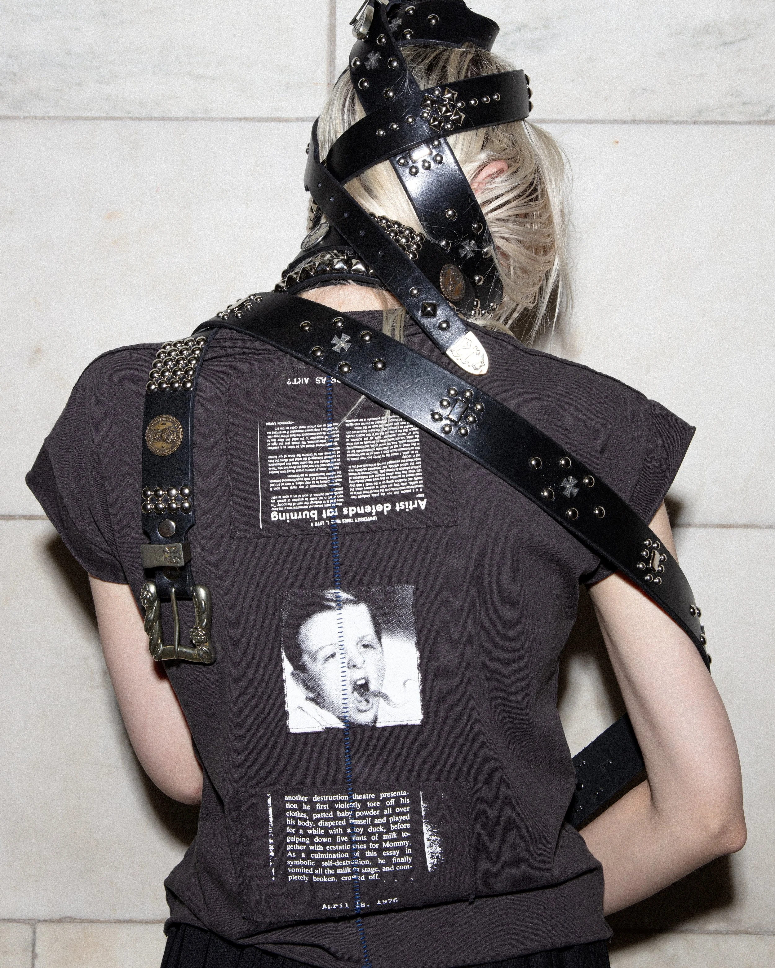 Person wearing a black T-shirt with a newspaper print graphic, accessorized with multiple black leather straps with studs and metal embellishments draped over shoulders and head.