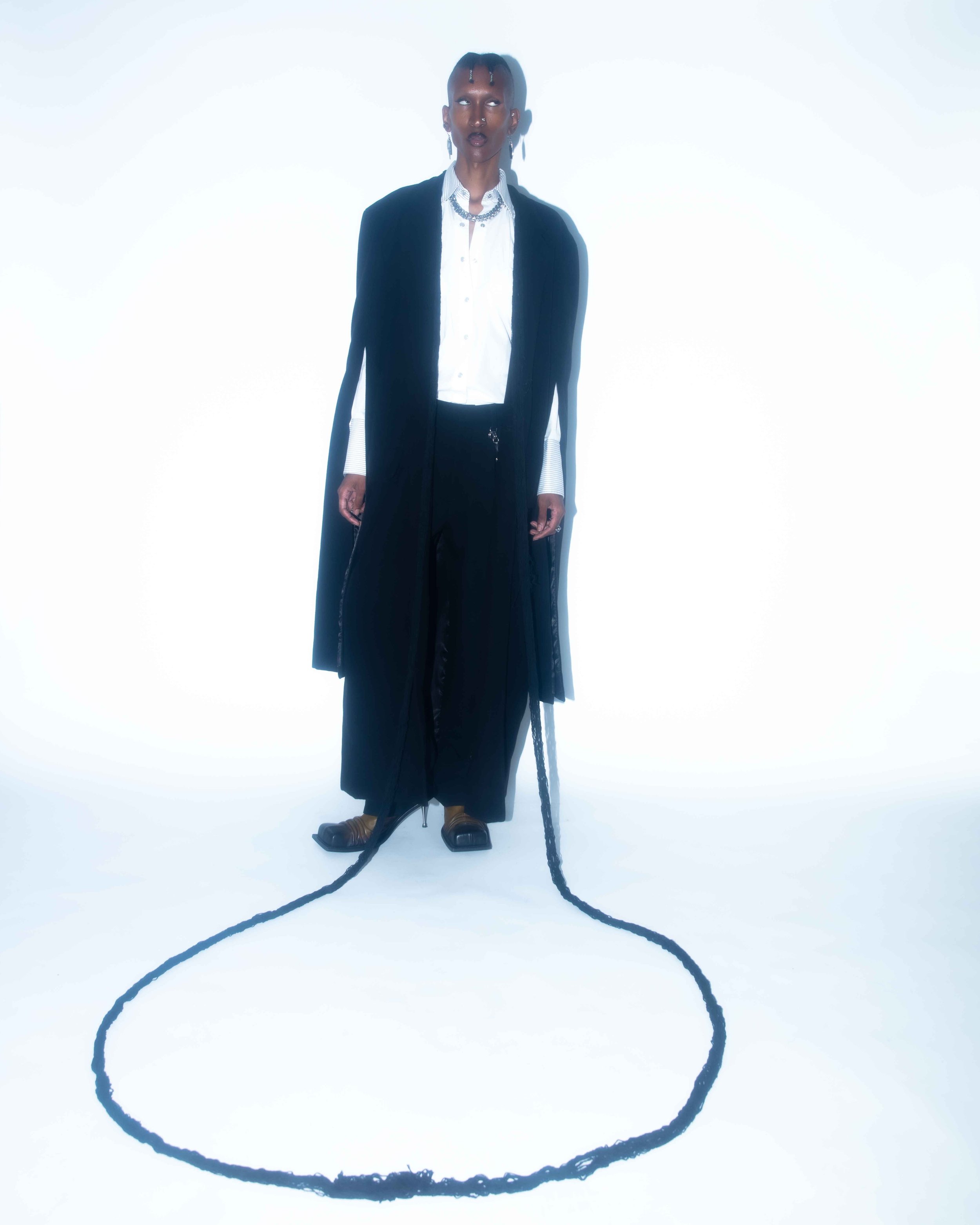Person with dark skin, wearing a long black coat, white shirt, black pants, and gold footwear, standing on a white background with a black rope on the ground.