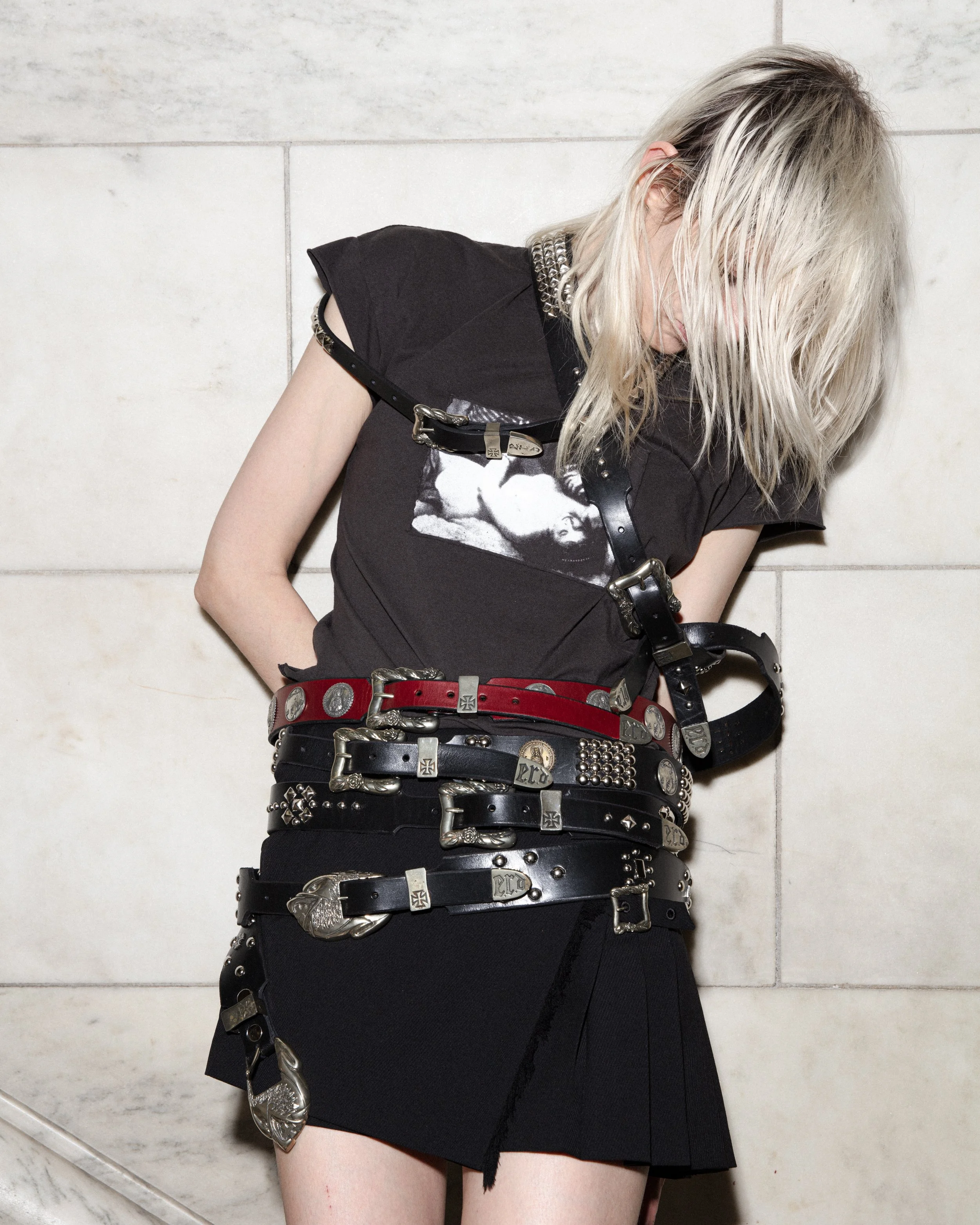 A woman with platinum blonde hair wearing a black T-shirt with a graphic print, multiple belts with metal decorations, and a black skirt, standing against a stone wall.
