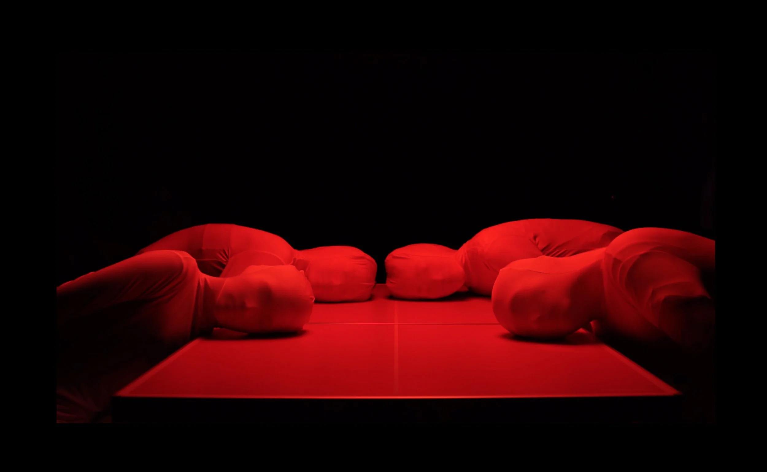 A person lying down on a red-lit ping pong table, covered entirely with a white sheet, in a dark room.
