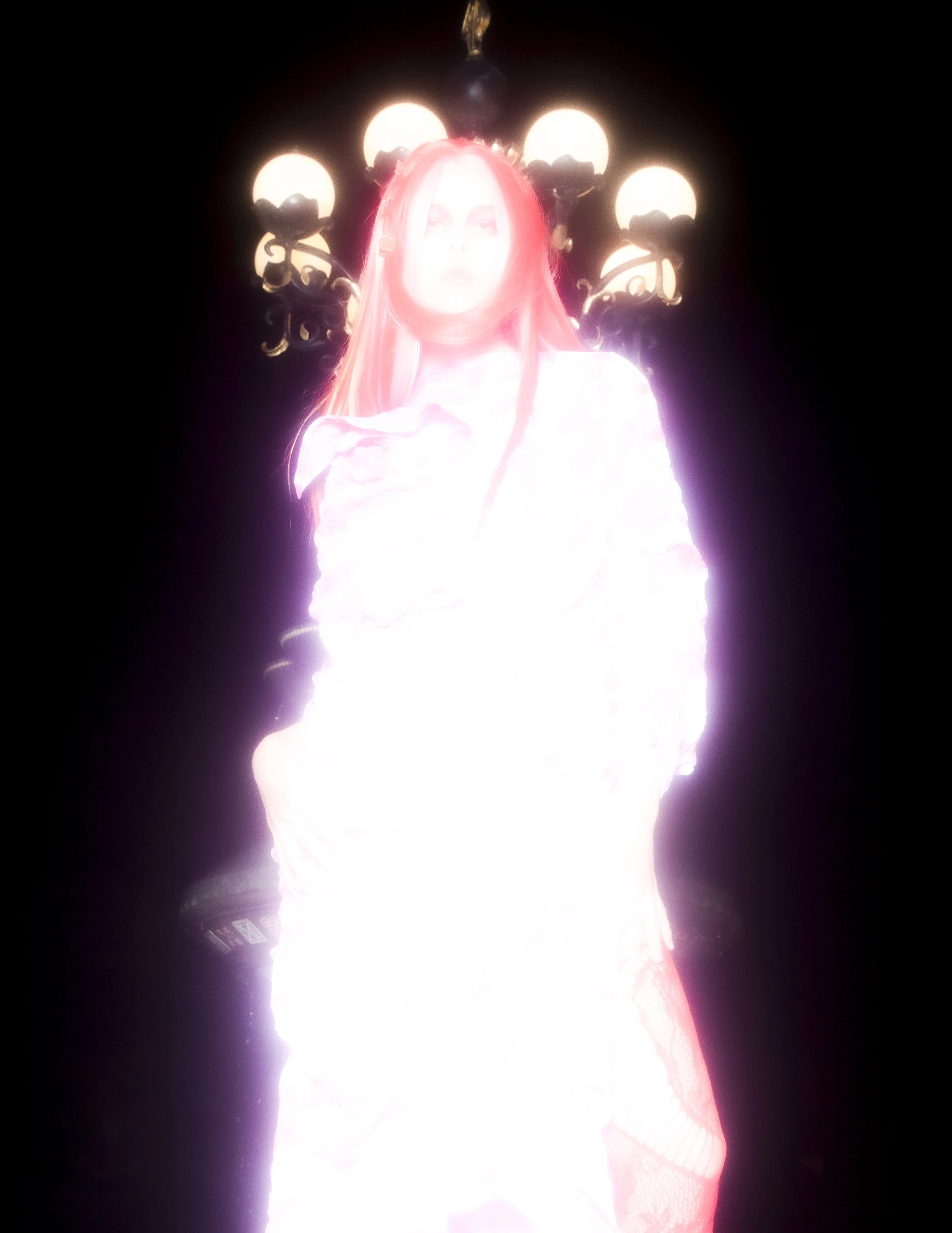 A person with long red hair standing in front of a chandelier with five glowing bulbs, emitting a bright light that overexposes the image and obscures facial features.