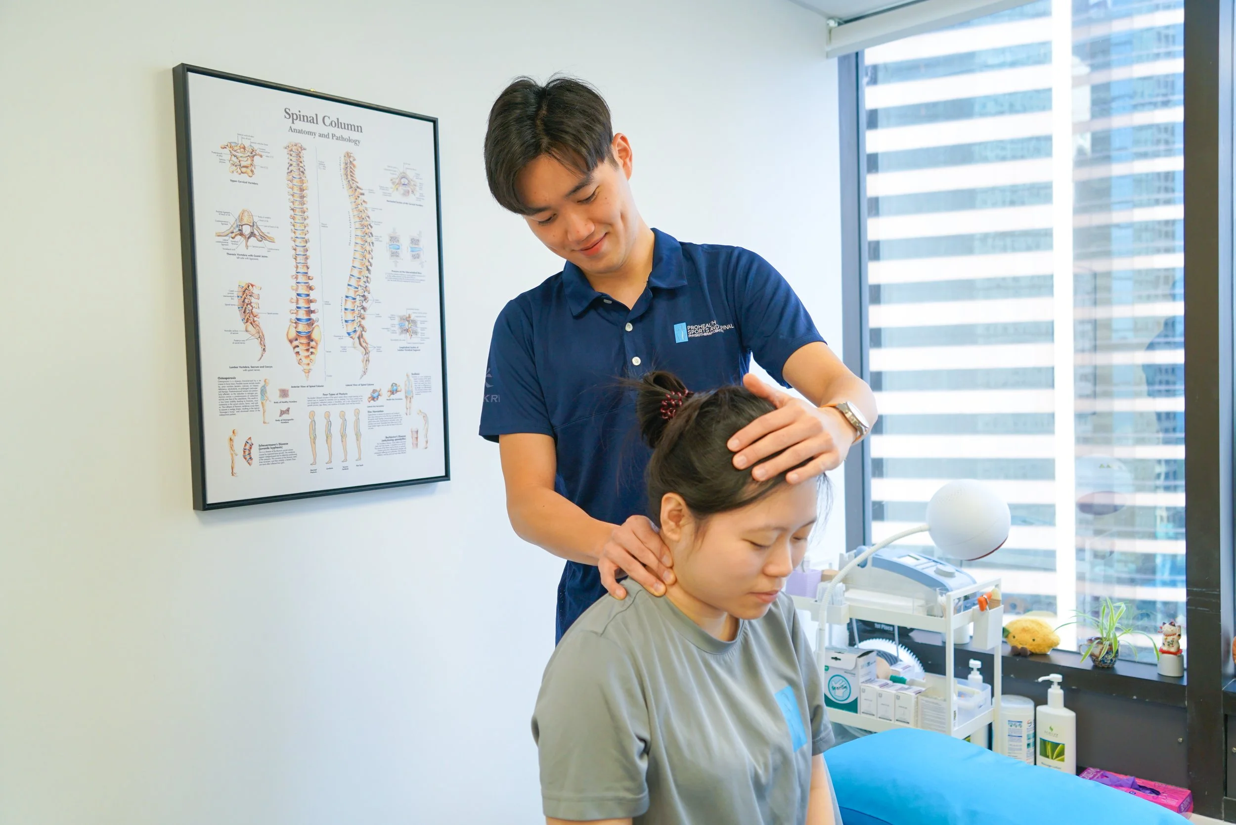 Chiropractor adjusting patient's neck in a medical office with a spinal column diagram on the wall.