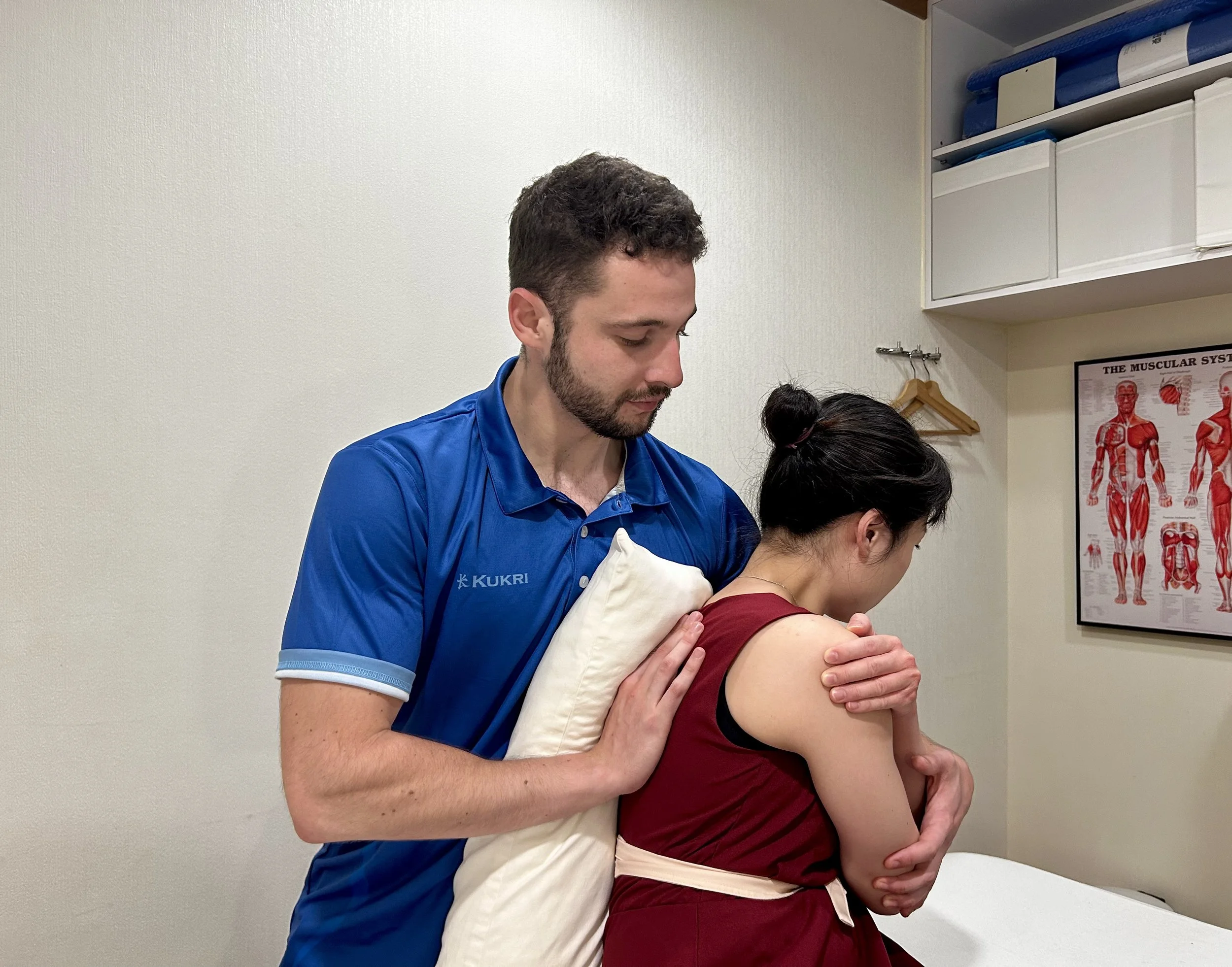 A massage therapist performs a seated shoulder massage on a woman in a clinic, with anatomical posters on the wall.
