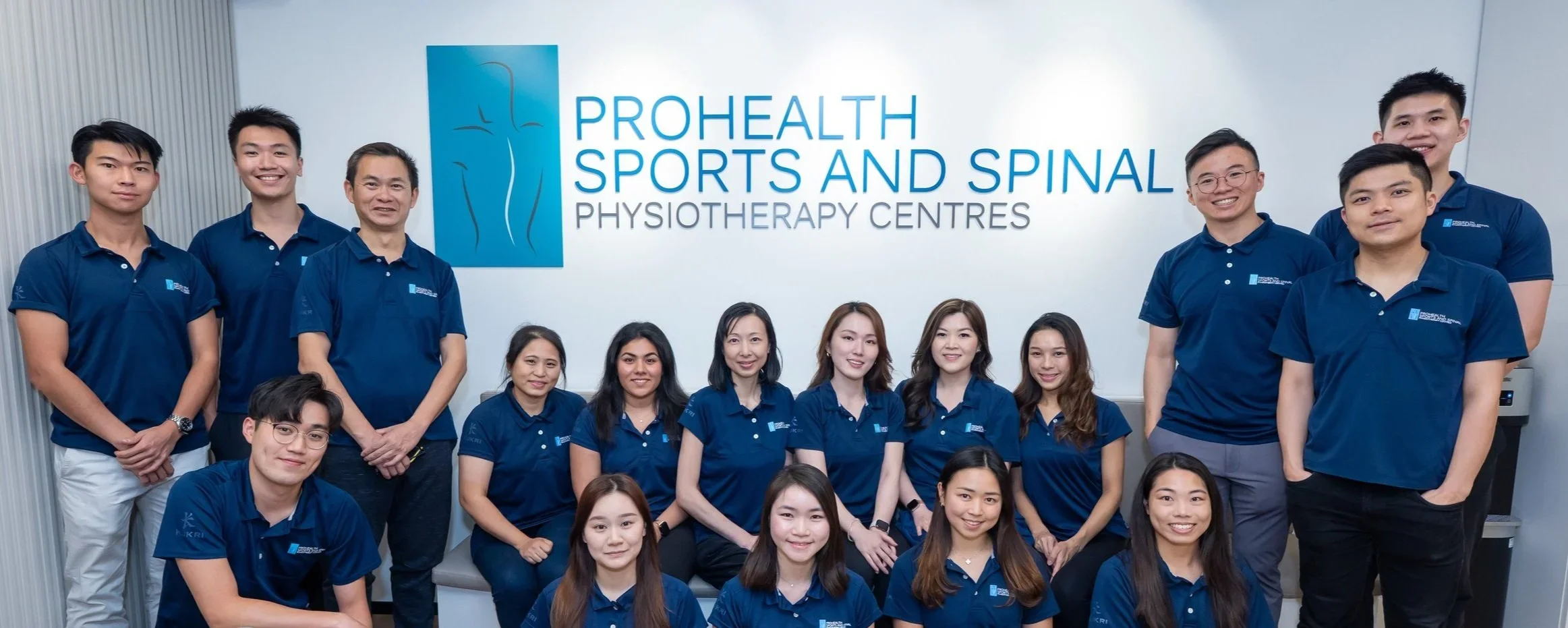 Group photo of staff members at ProHealth Sports and Spinal Physiotherapy Centres, all wearing matching blue polo shirts with the company's logo, standing and sitting in front of a wall with the company sign.