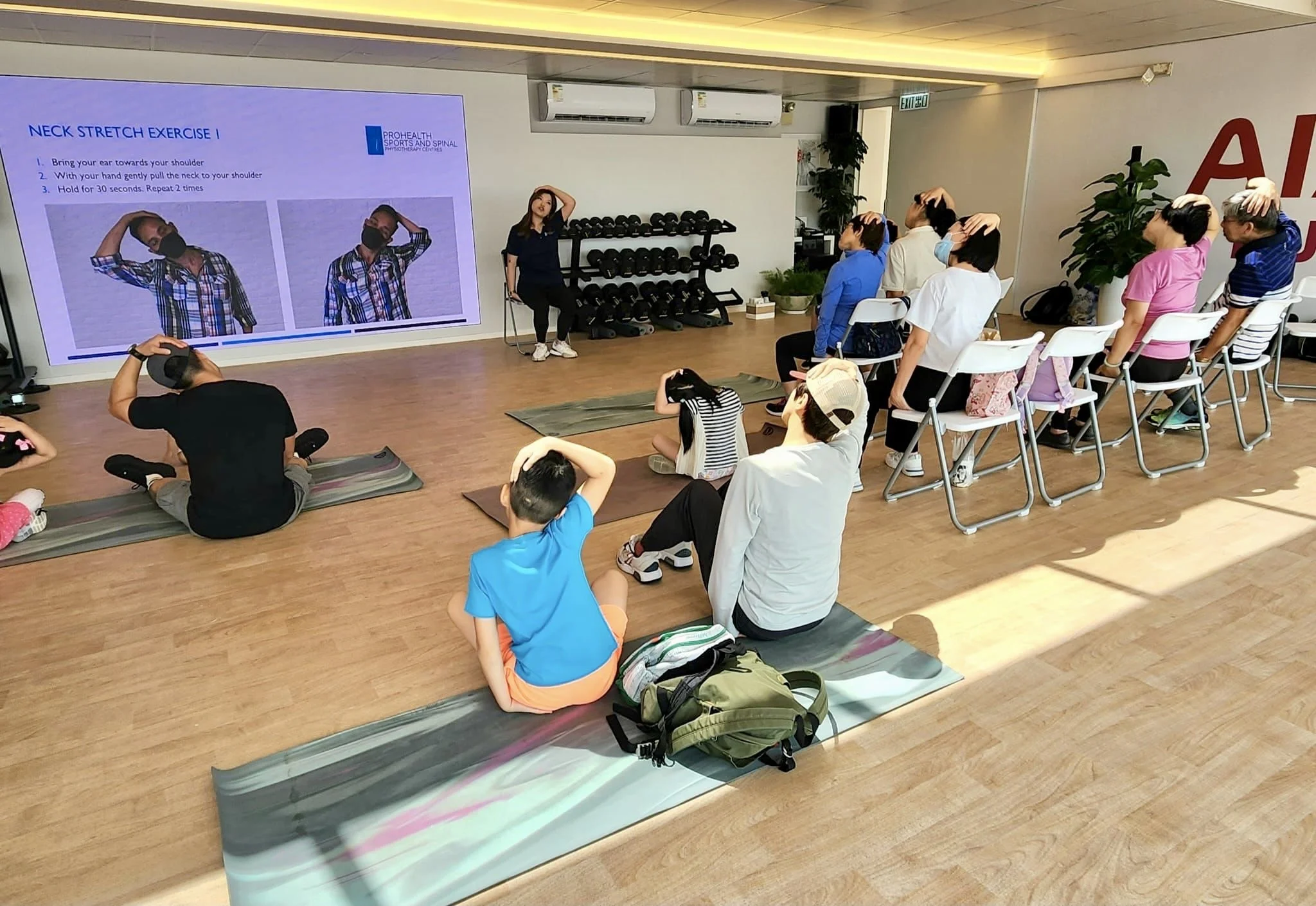 A group of children and adults participating in a stretching exercise class in a gym or wellness center. The instructor is sitting on a chair near a large screen displaying neck stretch instructions. Participants are sitting on mats and chairs, following along, with some children sitting on the floor and others on chairs. The room has wooden flooring, fitness equipment against the wall, and potted plants.