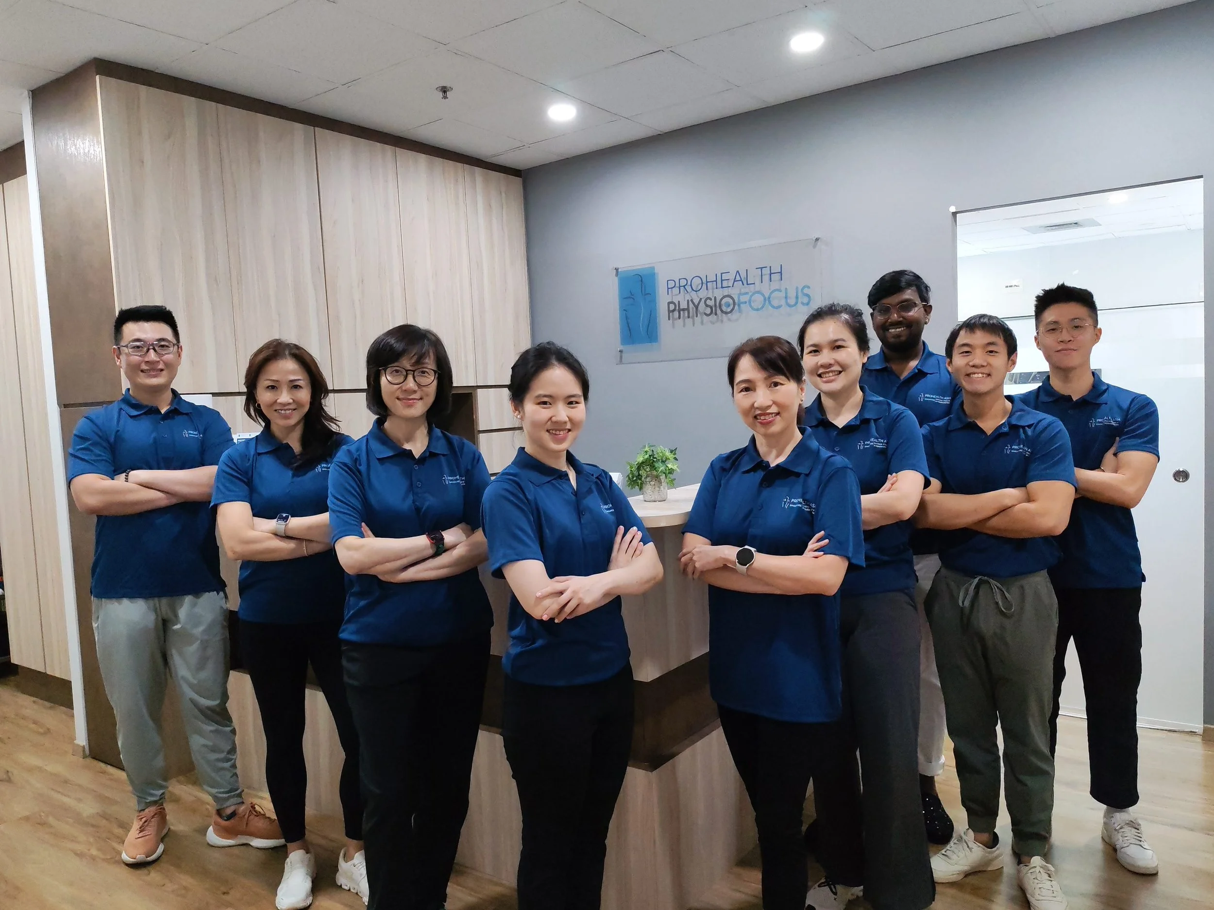 Group photo of nine diverse healthcare professionals wearing blue polo shirts standing with arms crossed in an office, with a sign that reads 'Prohealth Physiotherapy Focus' in the background.