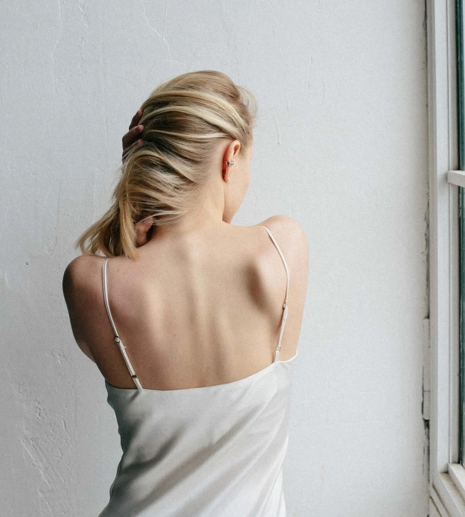 Blonde woman with balayage hair in a loose ponytail, facing a window in natural light, wearing a white silk slip dress — summer hair color AlbertColor NYC