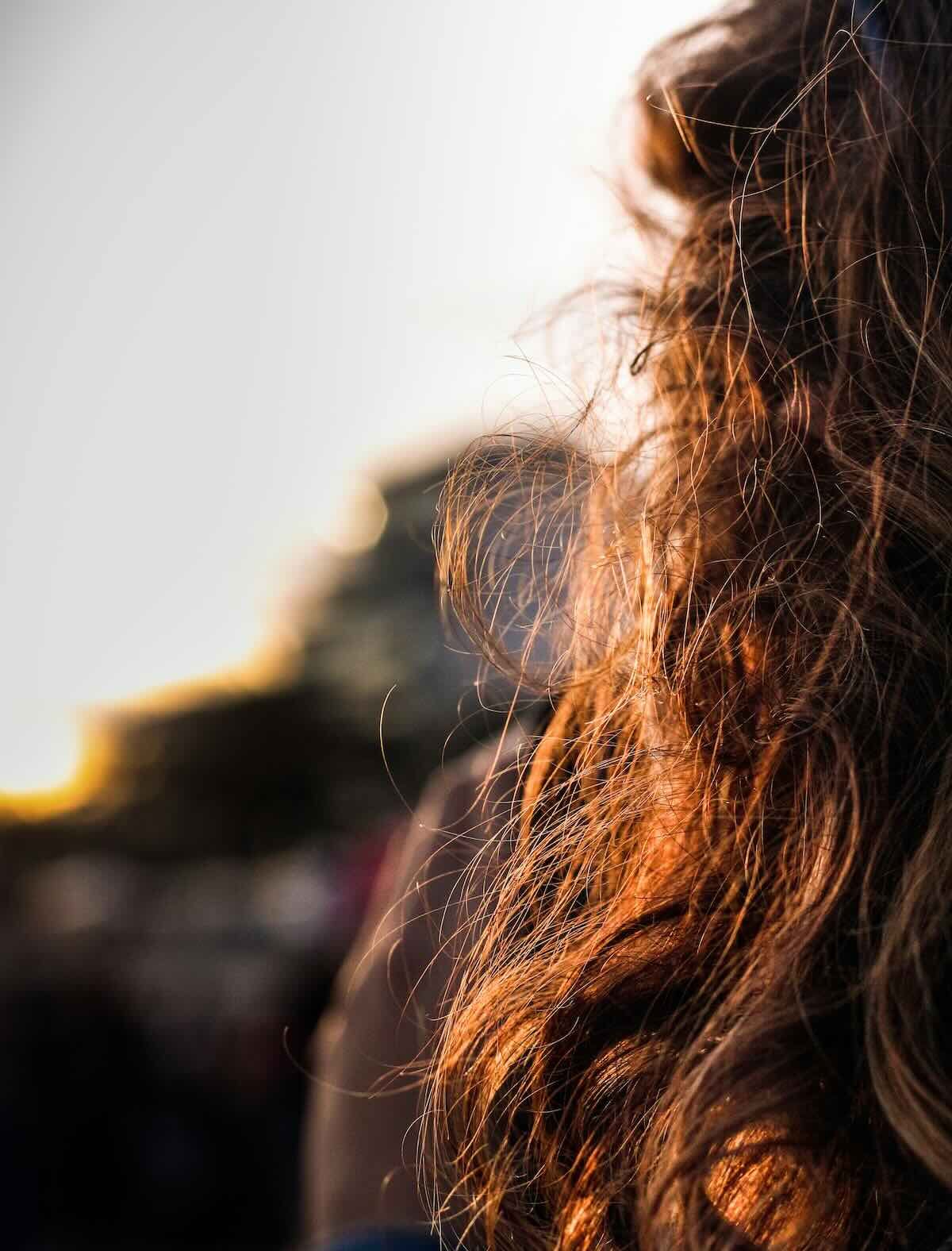 Backlit brown hair at golden hour, loose strands catching low evening sun against a pale sky, the light coming through the hair rather than onto it