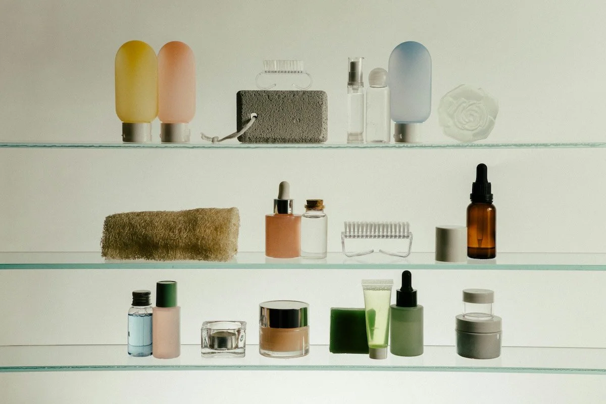 Three glass shelves against a pale wall, holding a spare arrangement of bottles, jars and brushes in soft yellow, peach, sage and amber, space left between them.