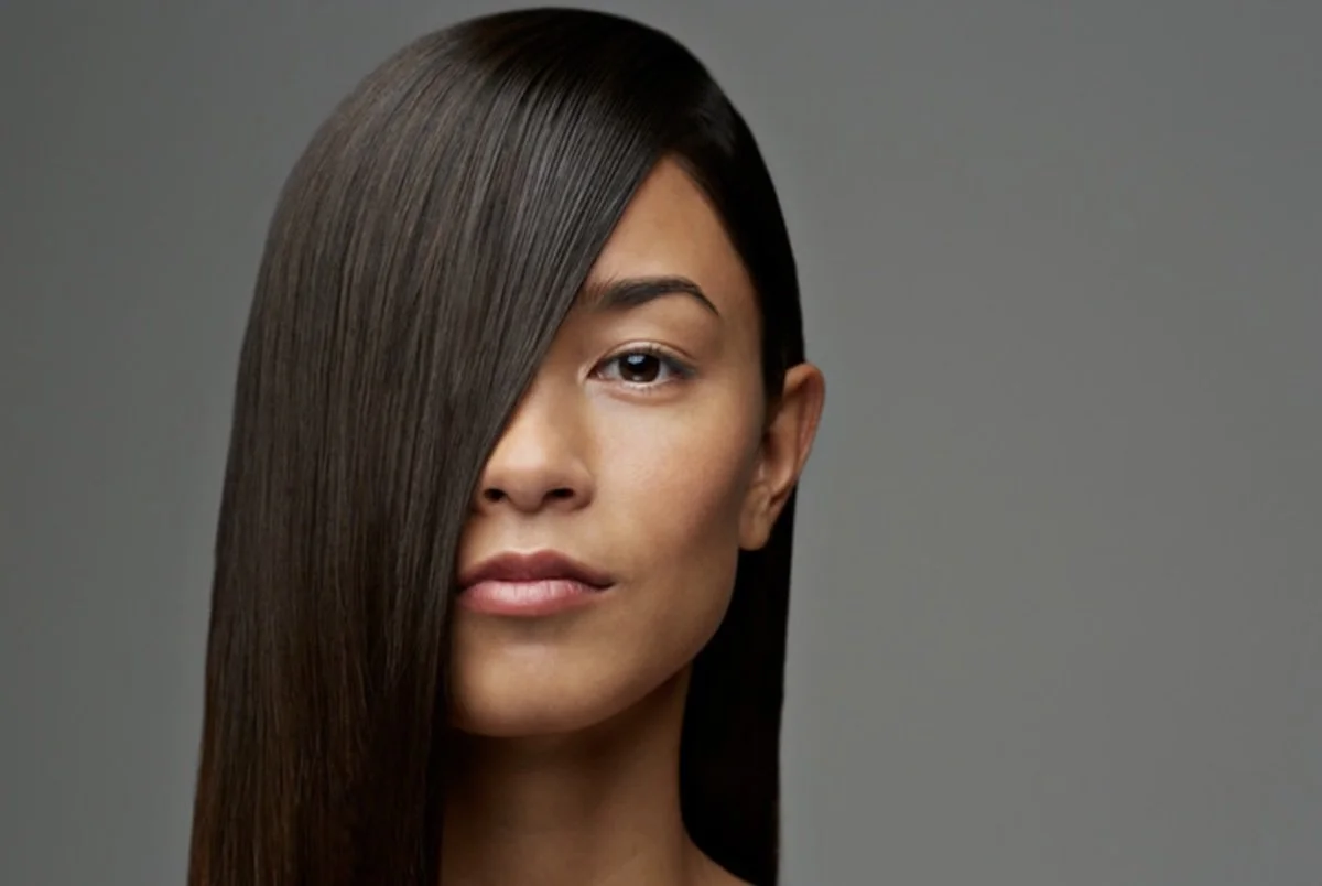 Dark brunette hair pressed poker-straight, one long panel swept diagonally across the face, the surface holding an even unbroken shine