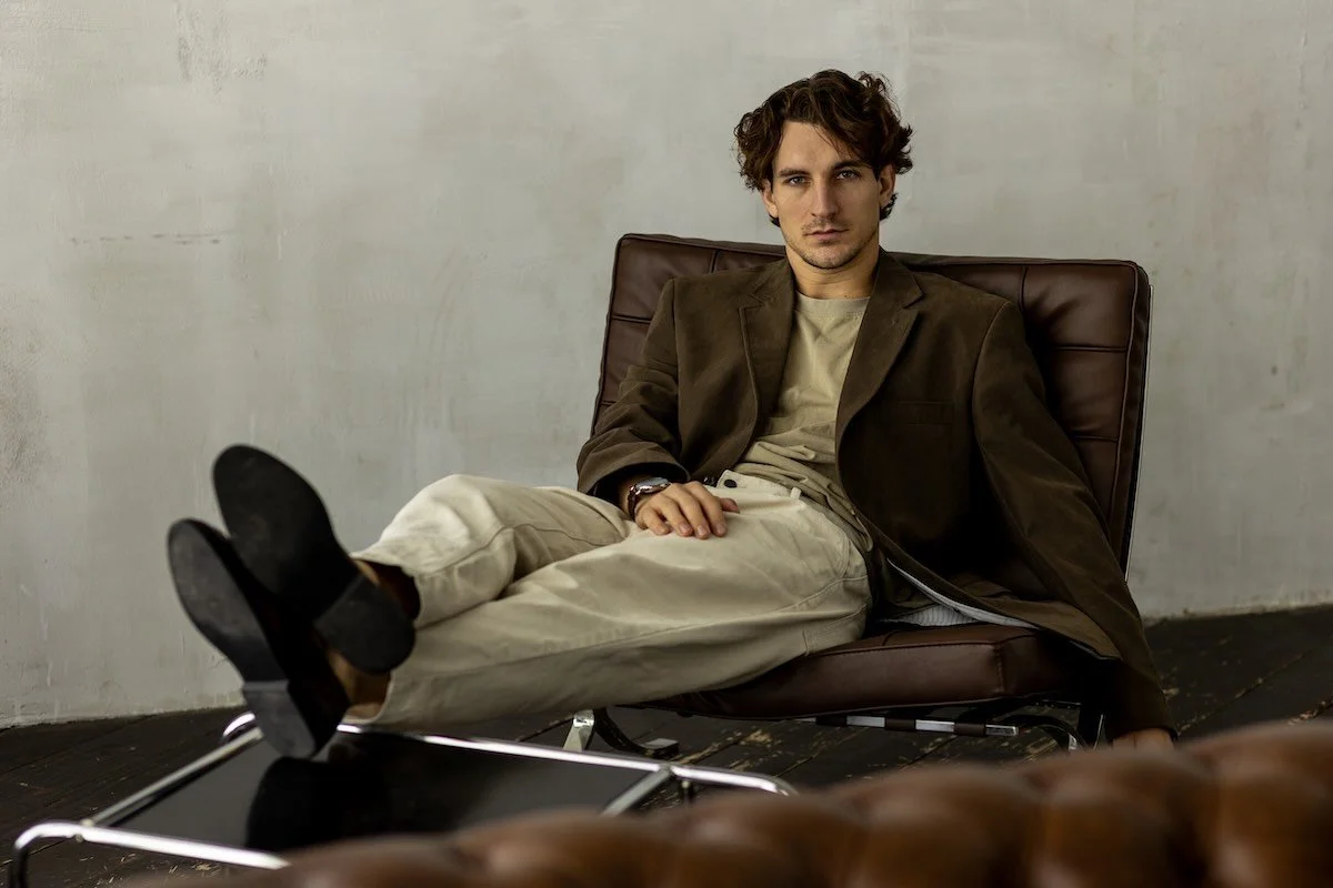 Man with dark wavy hair reclining in a brown leather chair, brown corduroy jacket over a beige tee and cream trousers, against a bare plaster wall
