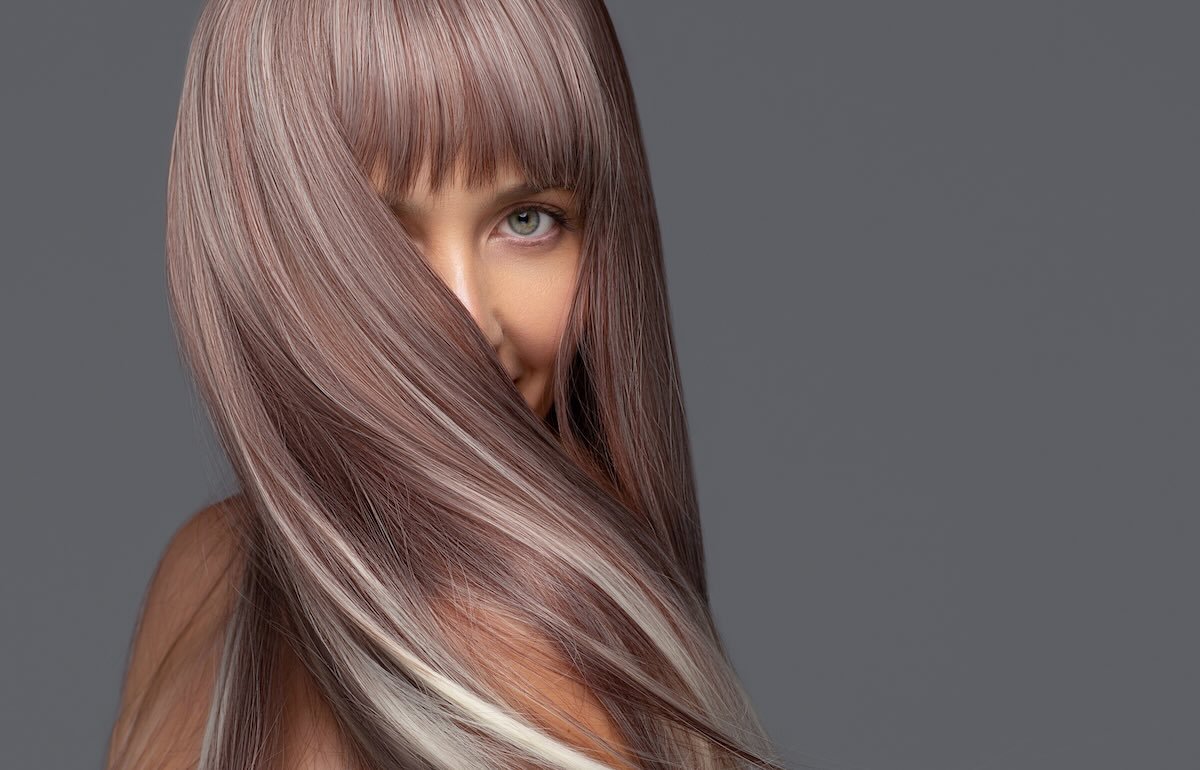 Long, silky, iridescent beige-blonde hair with pale blonde ribbons sweeps across a woman's face against a gray background, with one green eye showing through the strands.