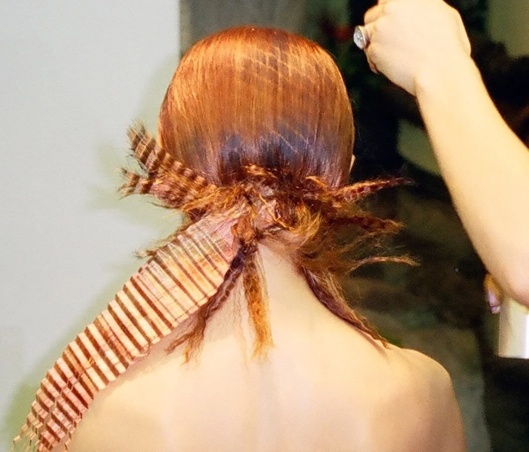 Back view of sleek copper hair gathered at the nape, crimped and pleated copper sections fanning over bare shoulders as a raised hand mists spray