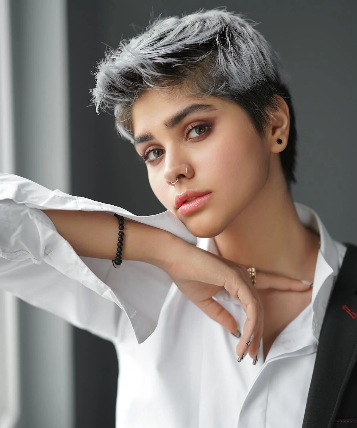 Woman with a short silver pixie cut over near-black sides and roots, in a white shirt and black blazer, chin resting on a raised forearm by a window
