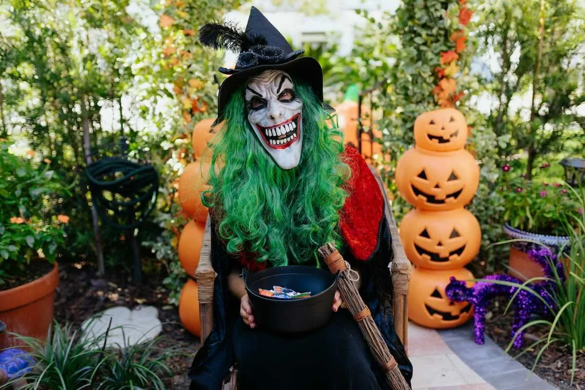 Person in a grinning witch mask, long curly green wig and black pointed hat, seated in a wicker chair holding a cauldron of candy beside stacked jack-o’-lanterns
